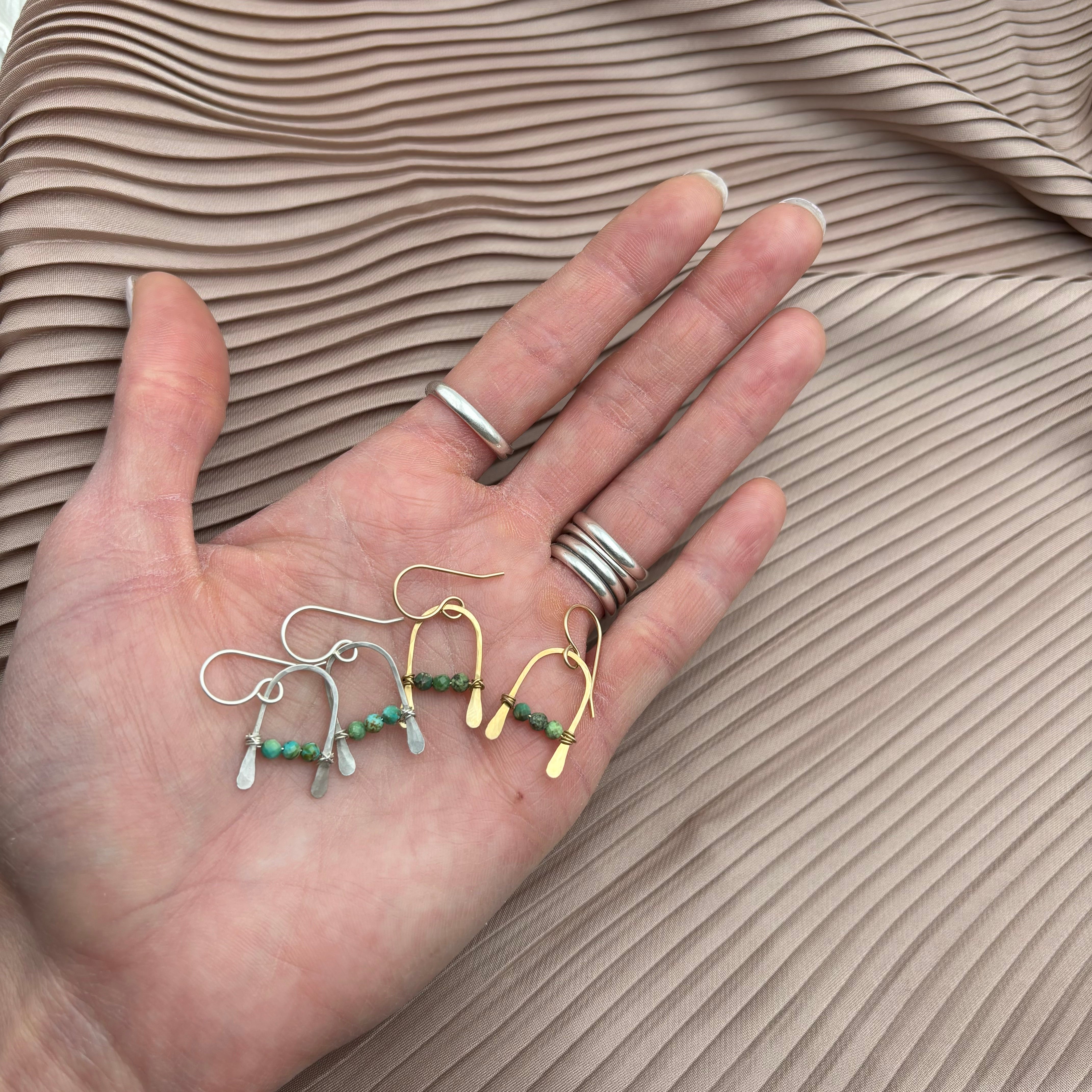 A hand with silver rings holds three pairs of Fluff Hardware Bitty Earrings—handmade with sterling silver charms and turquoise beads—displayed on pleated beige fabric.