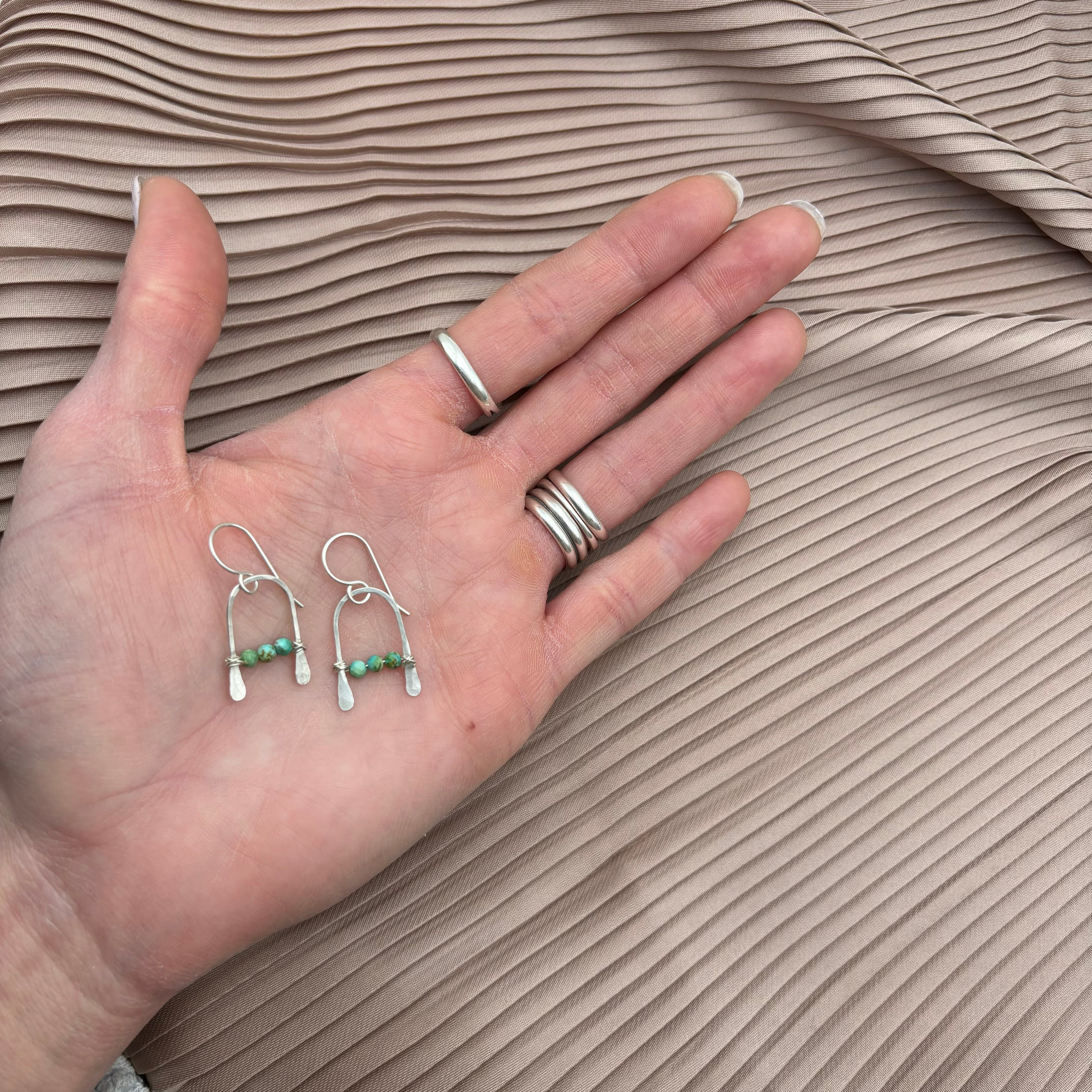 A hand with silver rings holds Fluff Hardwares Bitty Earrings—sterling silver wire earrings with small green beads—set against pleated light beige fabric.