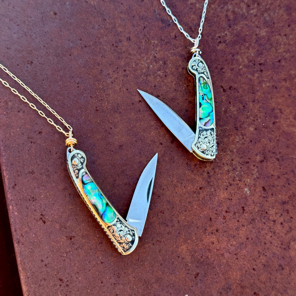 Two Lennox Necklaces by Fluff Hardware, featuring ornate utility pendant pocket knives with abalone inlays and silver detailing, hang from delicate chains; each blade is open and displayed on a brown textured surface.