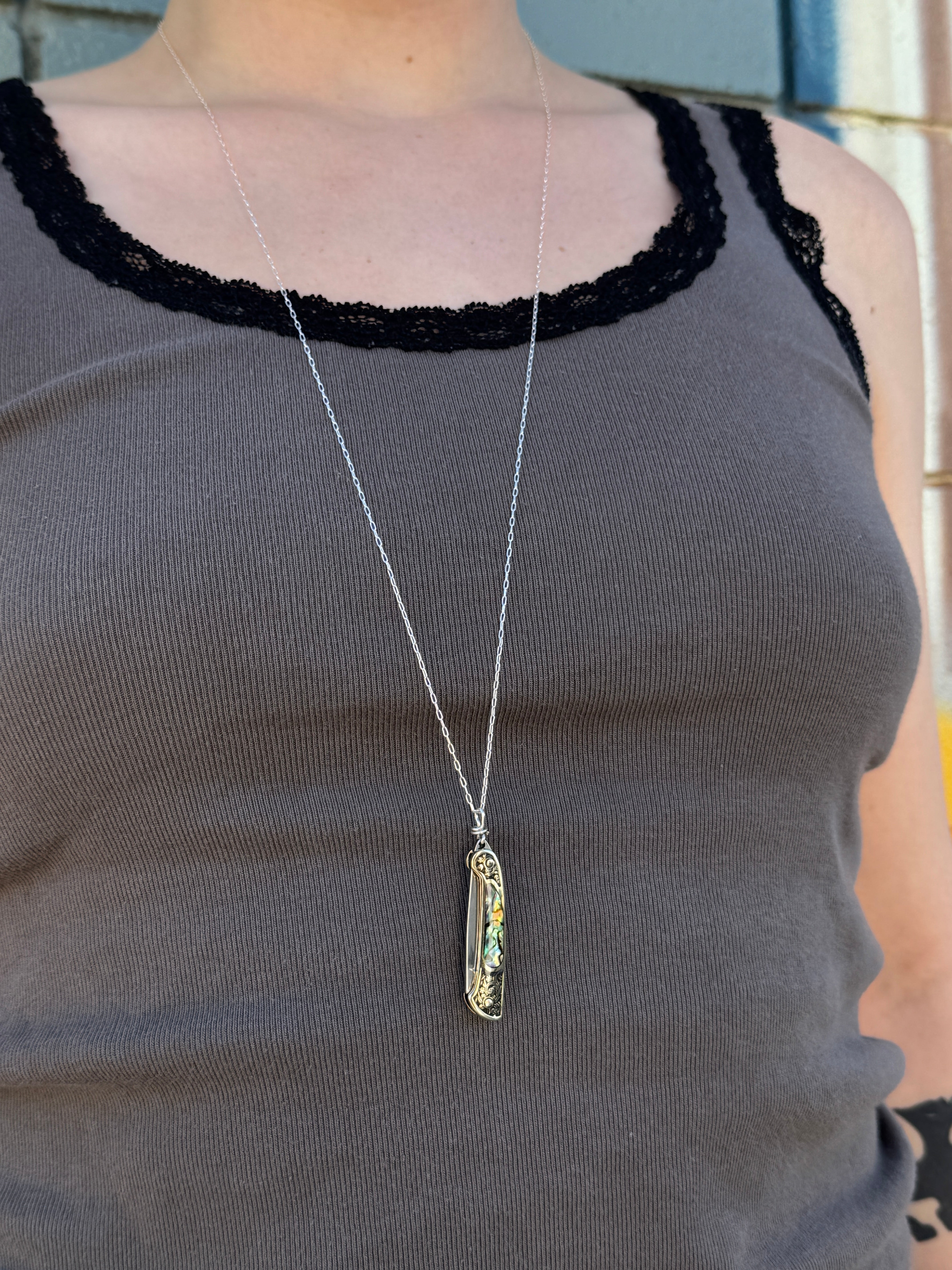 A person in a gray tank top with black lace trim is shown from shoulders to waist, wearing the Fluff Hardware Lennox Necklace—a long silver utility pendant with an oval charm featuring colorful details.