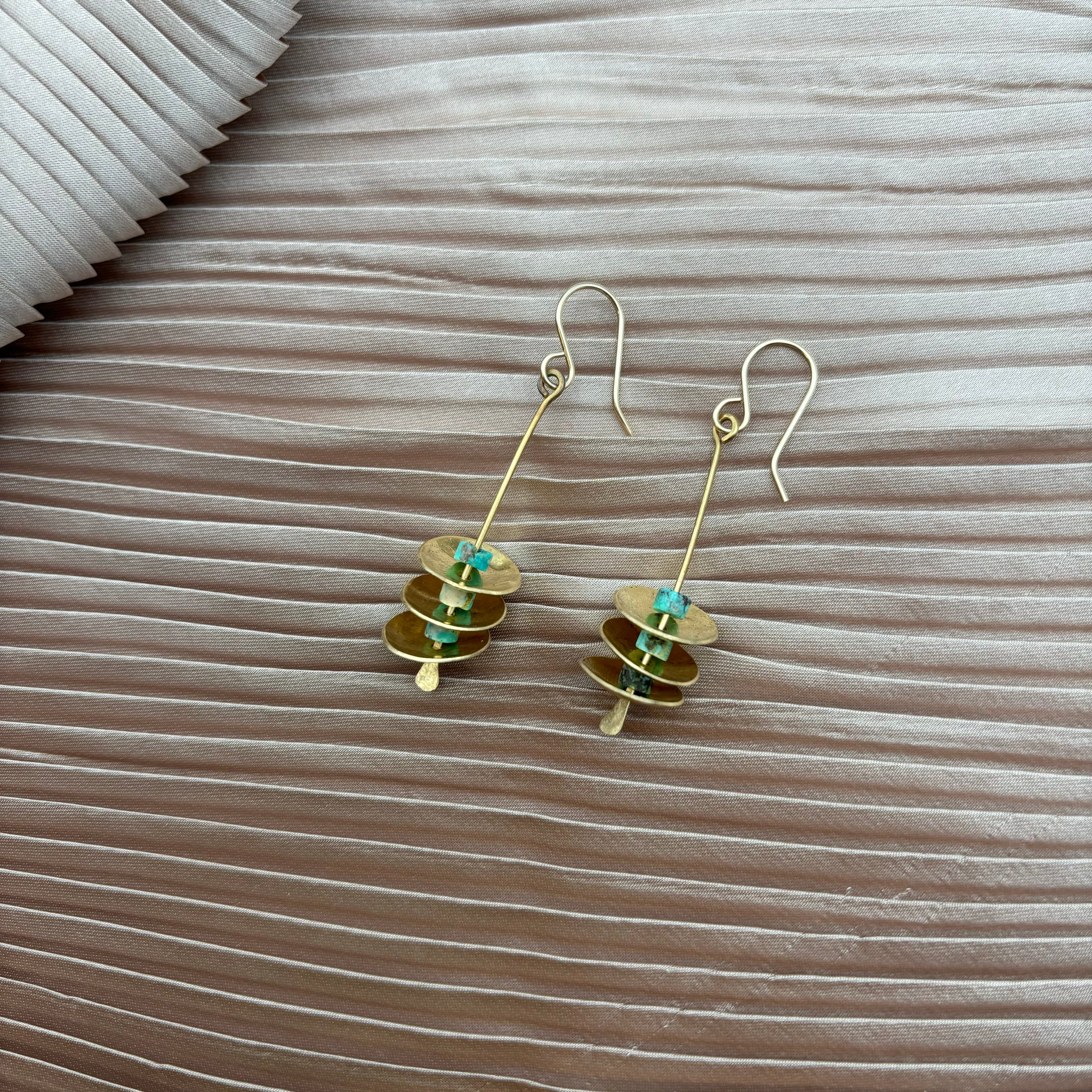 The Remains Earrings by Fluff Hardware feature stacked gold-filled round discs with small turquoise beads and hypoallergenic ear wires, displayed on textured light beige fabric.