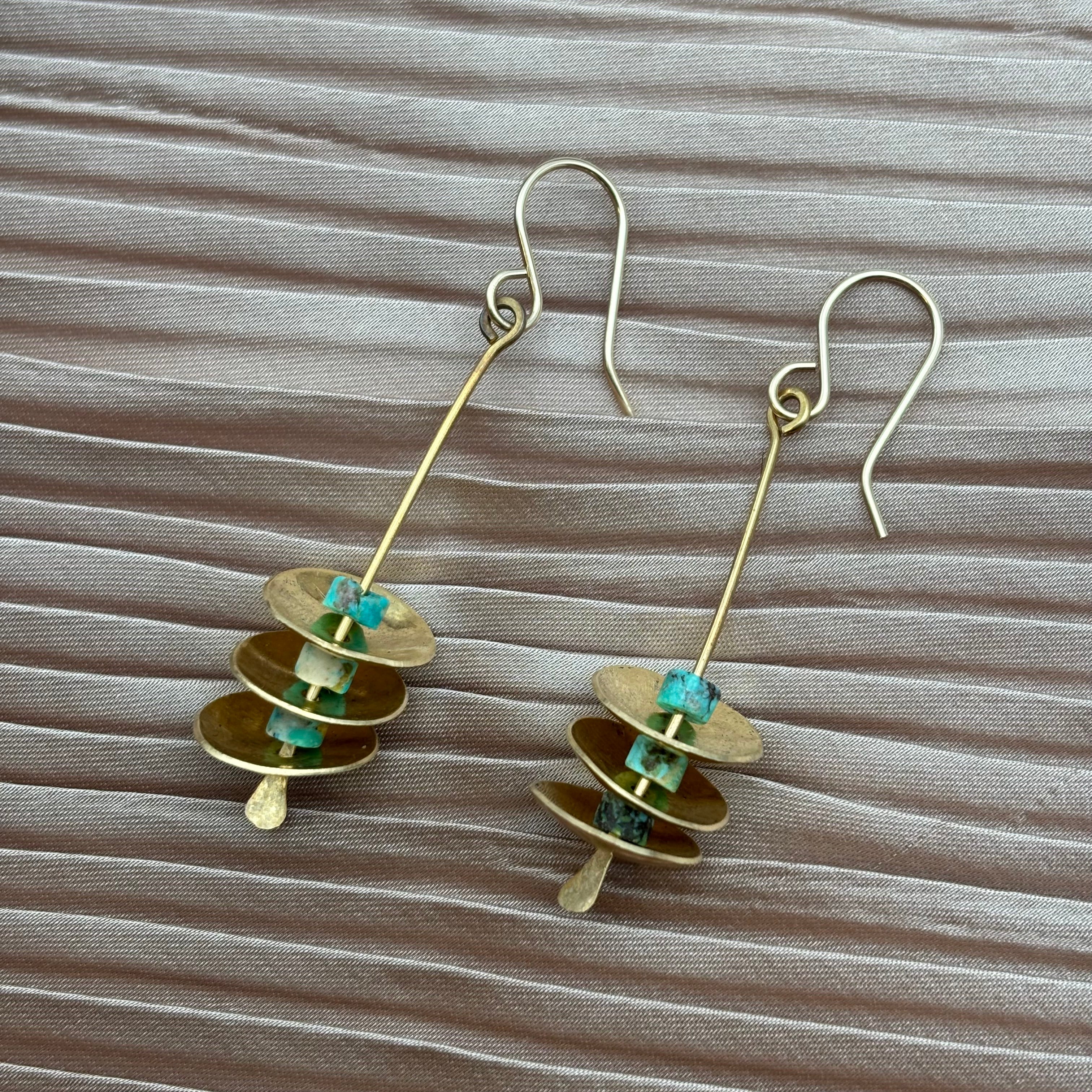 The Remains Earrings by Fluff Hardware feature gold dangle discs with turquoise bead accents and hypoallergenic ear wires, showcased on a light brown textured fabric background.