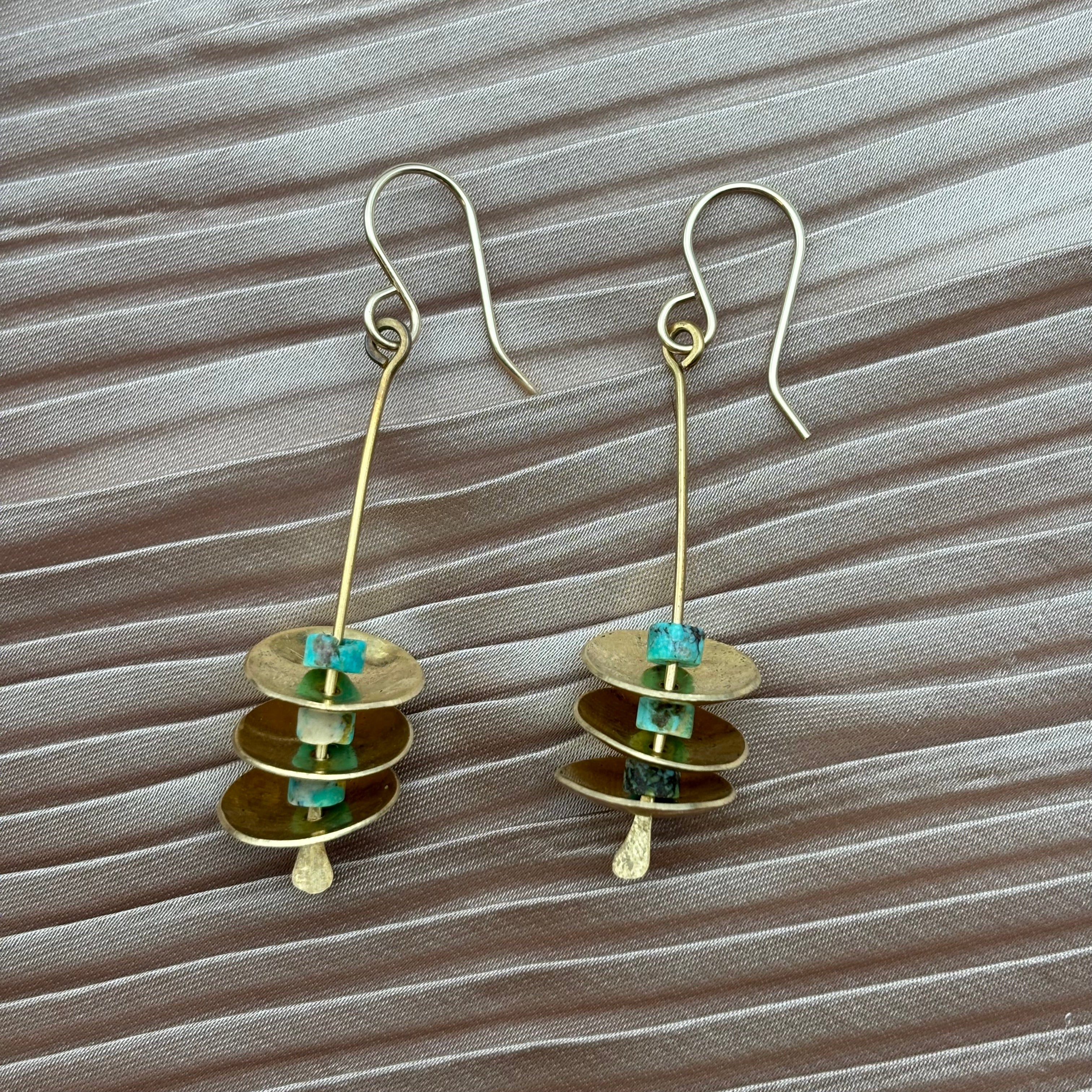 Remains Earrings by Fluff Hardware feature gold-filled hooks, slim vertical rods, and flat gold discs separated by turquoise beads, all displayed on textured light brown fabric.