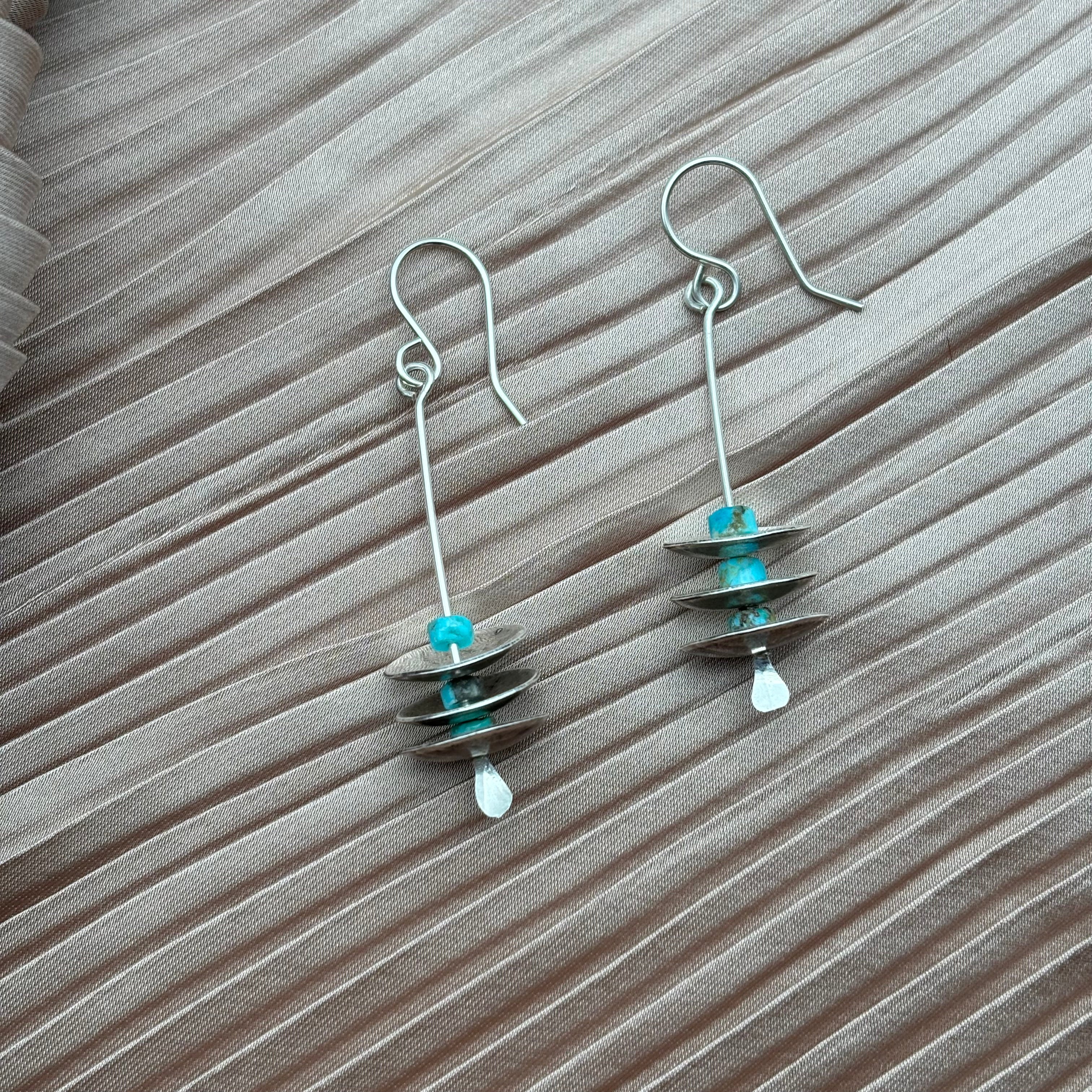 The Remains Earrings by Fluff Hardware are handmade dangle earrings with turquoise beads and silver discs on a silver wire and hypoallergenic ear wires, shown on a textured wooden surface.
