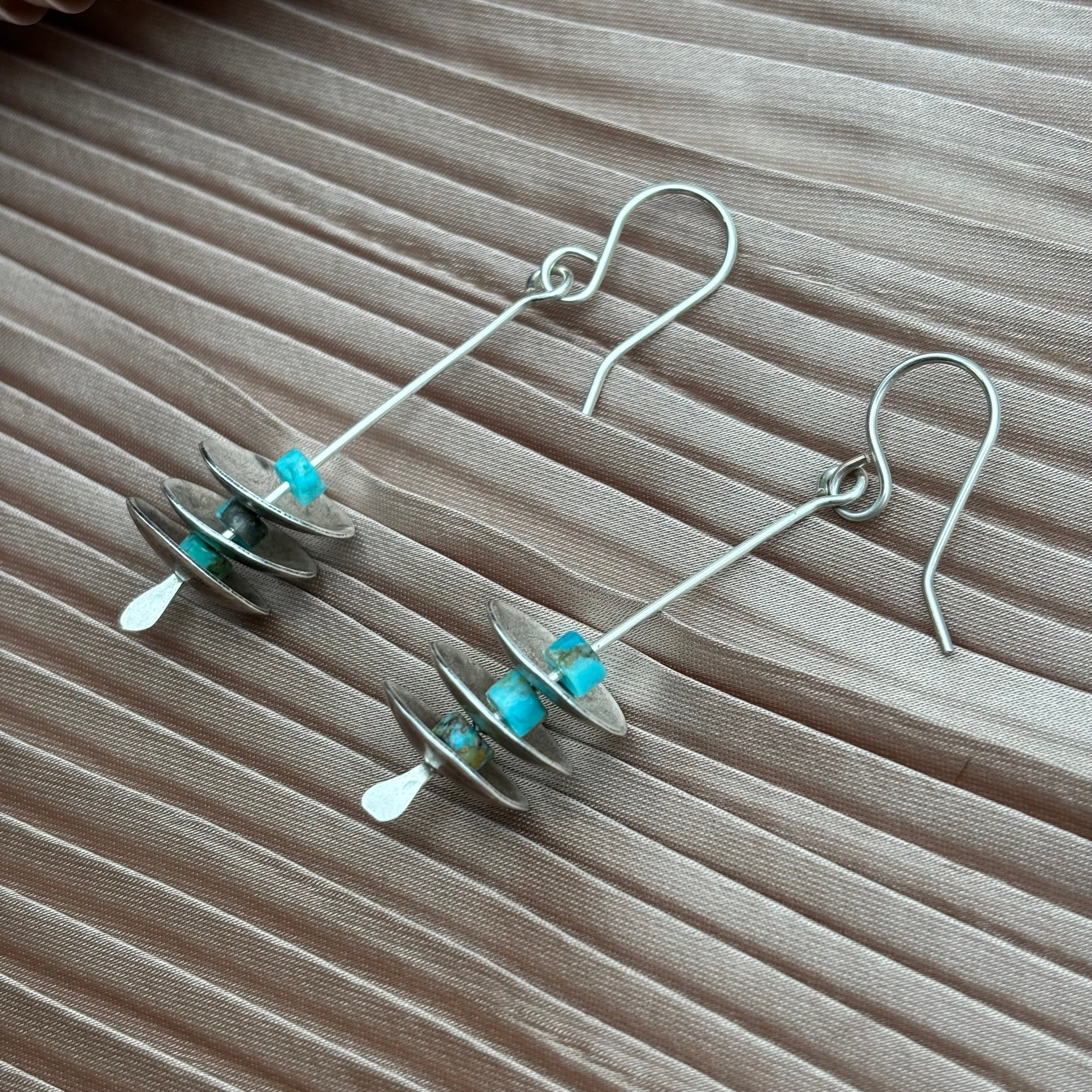 The Remains Earrings by Fluff Hardware are modern sterling silver dangle earrings with stacked metal discs and turquoise-blue beads, hypoallergenic ear wires, shown on taupe fabric.
