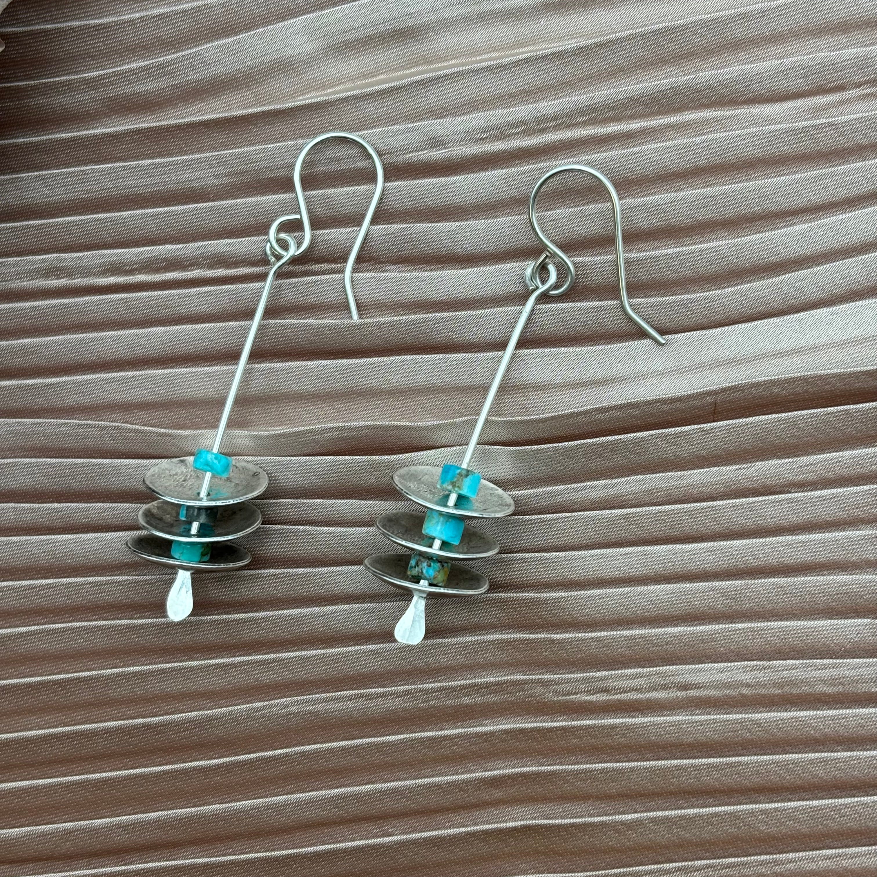 The Remains Earrings by Fluff Hardware are dangling sterling silver earrings with hypoallergenic ear wires, stacked silver discs, and turquoise beads, shown on beige pleated fabric.