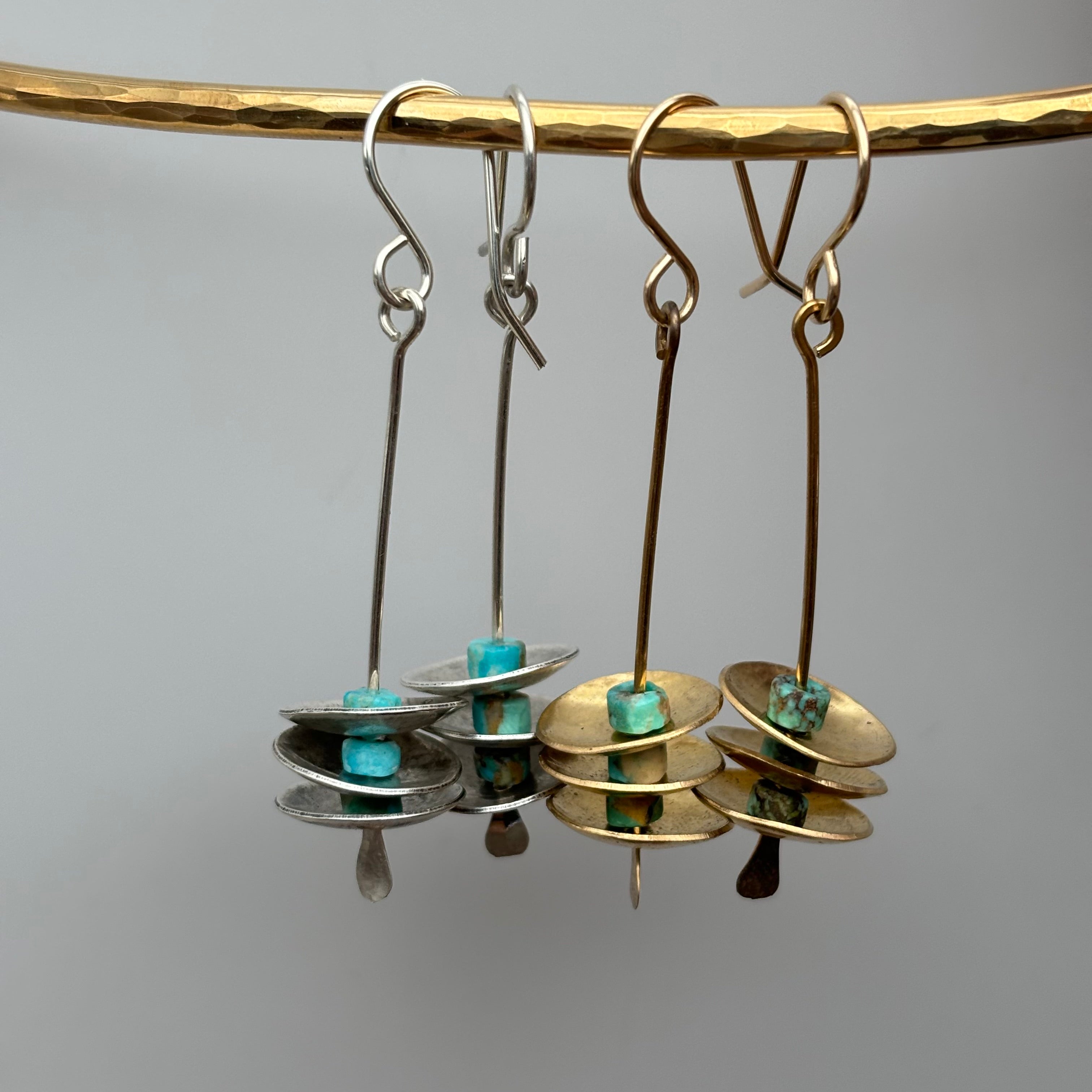 Four Remains Earrings by Fluff Hardware hang on a gold rod—two in sterling silver with turquoise, two in gold with green stones. Each earring has stacked circular discs, hypoallergenic ear wires, and a small drop, set against a plain gray background.