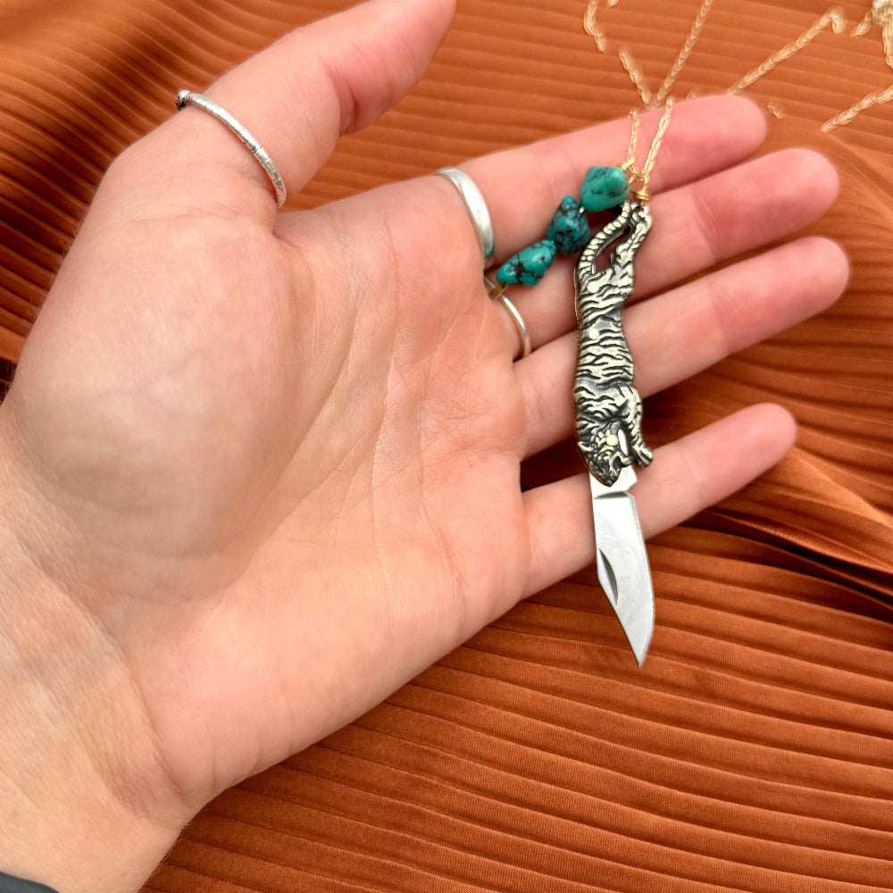 Hand holding a mini knife with a tiger shaped handle on a necklace chain with turquoise stones and with the blade out