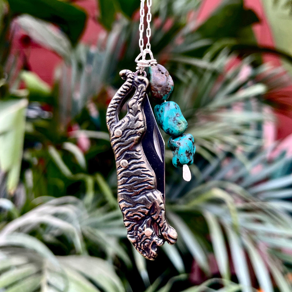 Tiger-shaped pocketknife pendant with turquoise stones on a chain against a green leafy background
