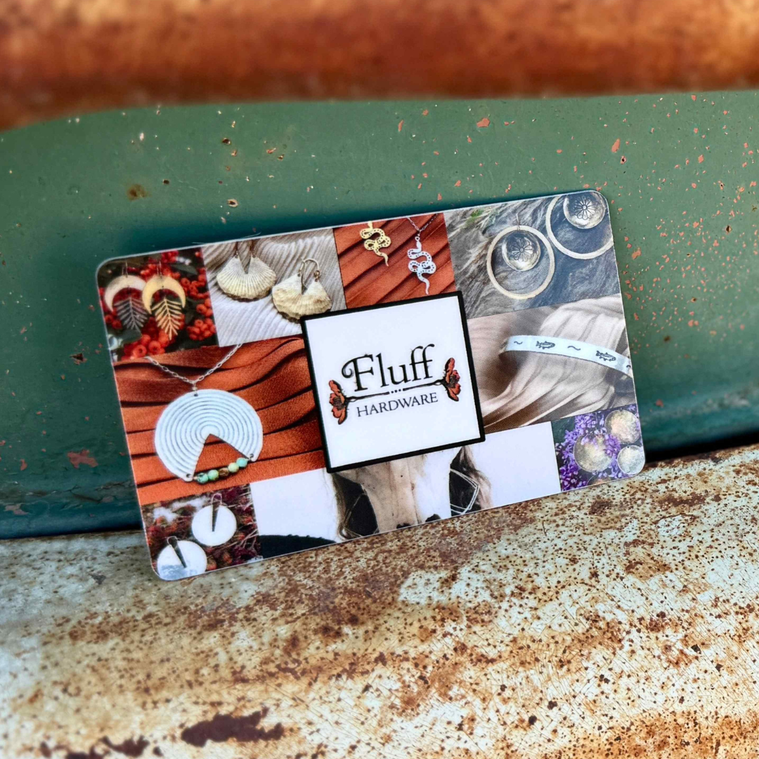 The Fluff Hardware Other Gift Card is redeemable online and features photos of handmade earrings, necklaces, and rings displayed on a rustic metal surface with a green and brown backdrop.