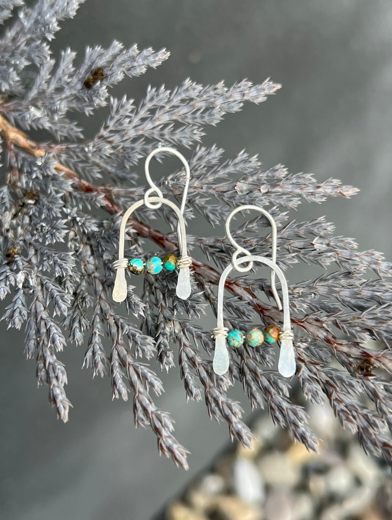 Bitty Earrings by Fluff Hardware: Handmade sterling silver wire earrings with teardrop white stones and small turquoise beads, shown hanging on a branch with silvery-gray foliage.