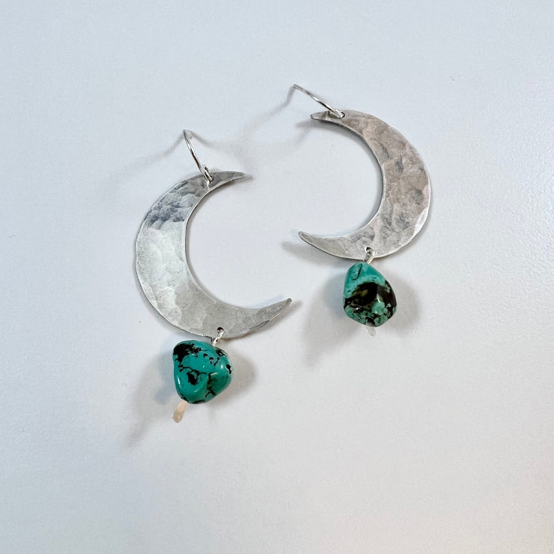 Fluff Hardware’s Turquoise Moon Earrings feature hammered silver moons with dangling turquoise stones. Hypoallergenic and displayed on a light background, these earrings add celestial charm to any look.