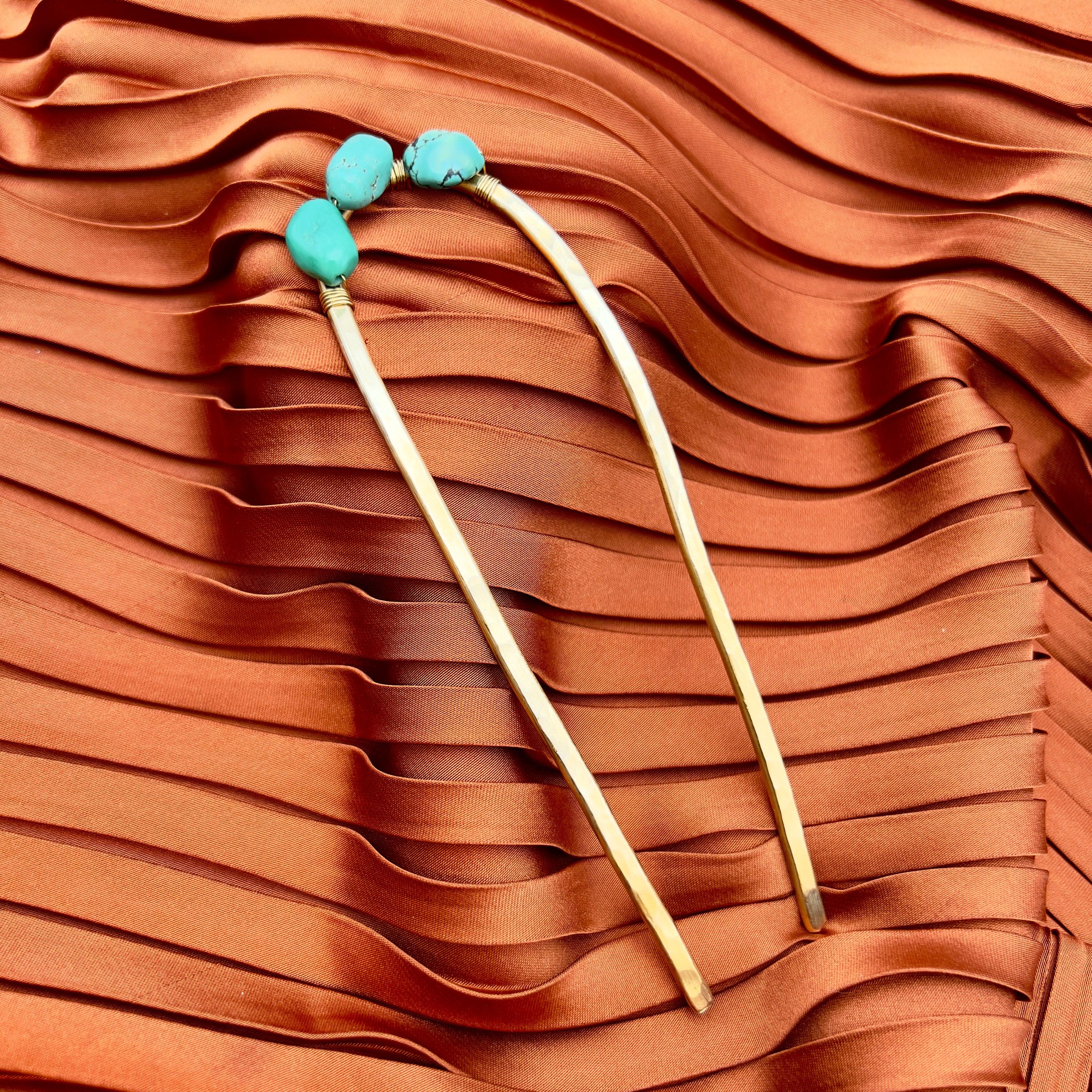 The Turquoise Hair Fork by Fluff Hardware features gold sticks with turquoise beads, displayed on rust-colored satin—perfect for adding elegance to your updo or trying a hair fork tutorial.