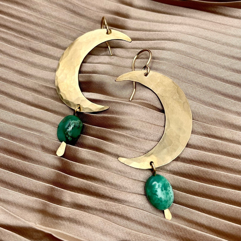 Fluff Hardwares Turquoise Moon Earrings—large, hammered brass moons with turquoise stones—are displayed on pleated beige satin fabric.