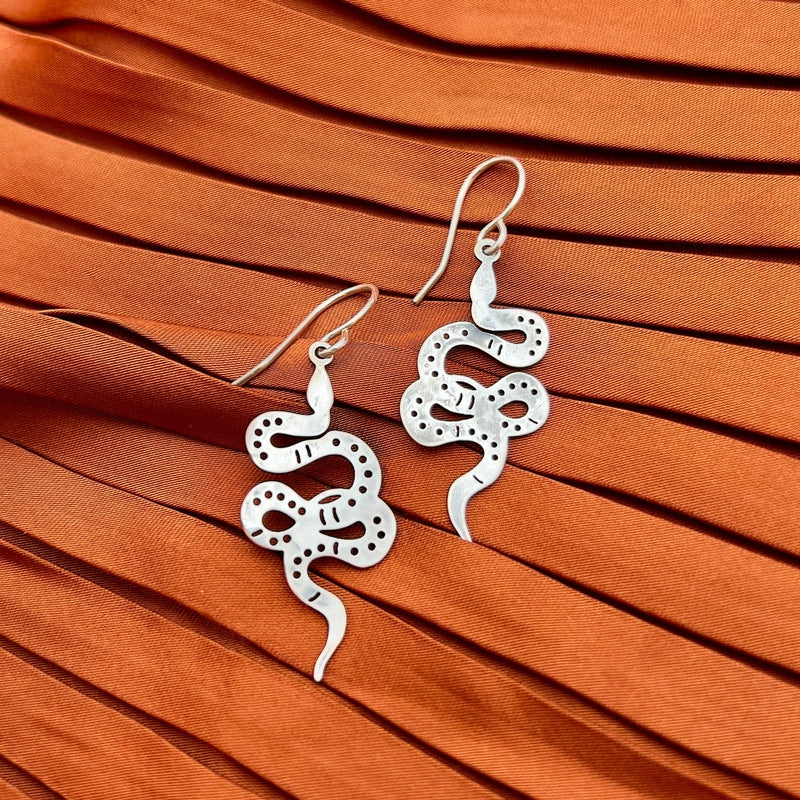 The Slither II Earrings by Fluff Hardware—silver, snake-shaped with dotted patterns and delicate pendants—are shown on pleated rust-colored fabric.