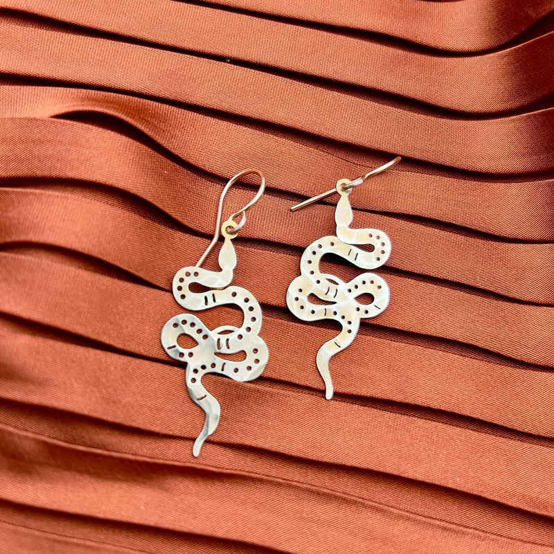 The Slither II Earrings by Fluff Hardware, featuring metallic snake shapes with cutout and dot details and 14k gold filled accents, are displayed on textured rust-colored pleated fabric.