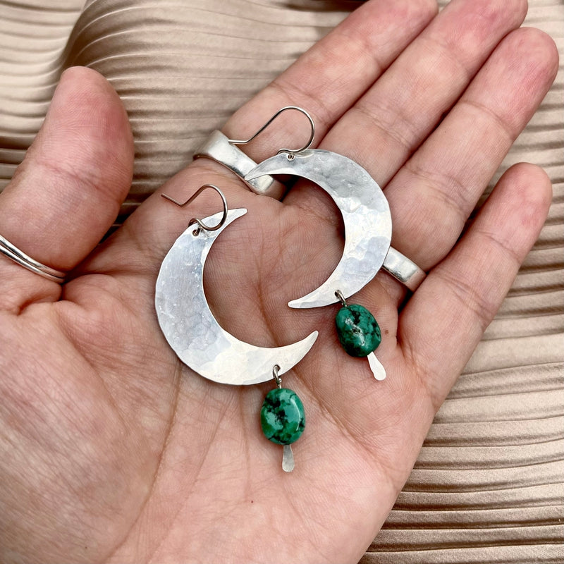 A hand displays Fluff Hardware’s Turquoise Moon Earrings—crescent-shaped with green marbled turquoise stone dangles—set on pleated beige fabric, perfect for anyone seeking distinctive, hypoallergenic jewelry.