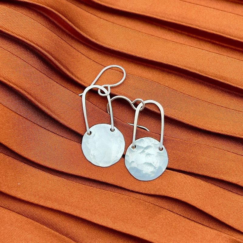 The Fallen Moon Earrings by Fluff Hardware—hammered silver disc earrings with hook wires, ideal as hypoallergenic jewelry—are displayed on pleated orange fabric.