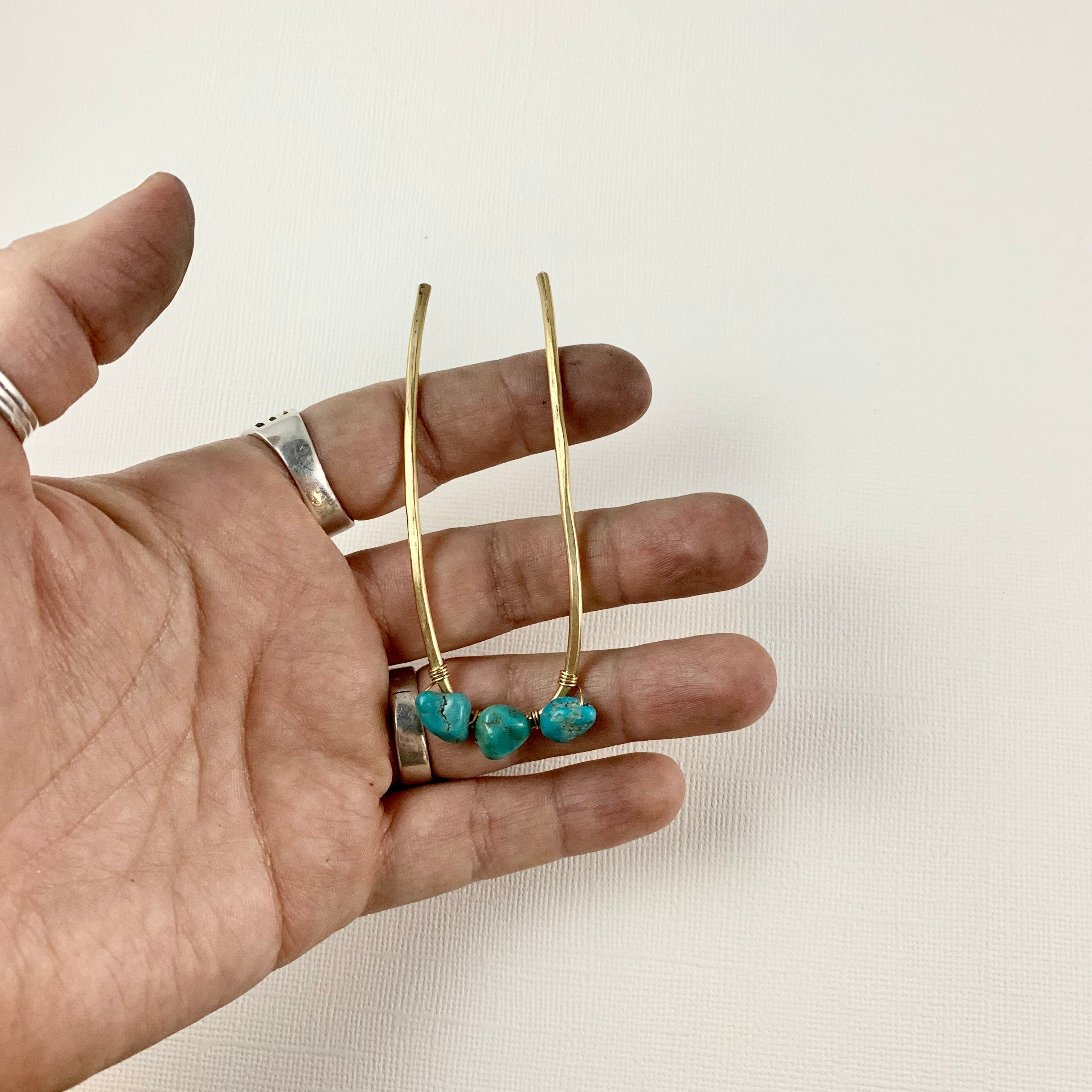 A hand holds the Fluff Hardware Turquoise Hair Fork, featuring a long, curved gold design with turquoise beads. The model wears silver rings against a plain background—perfect inspiration for styling your hair with this elegant accessory.