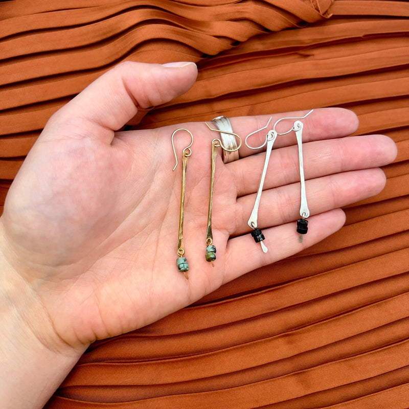 A hand displays two pairs of Fluff Hardwares Spark Earrings—one sterling silver with black and white beads, the other gold with green beads—against orange pleated fabric. Both hypoallergenic designs combine elegance and comfort.