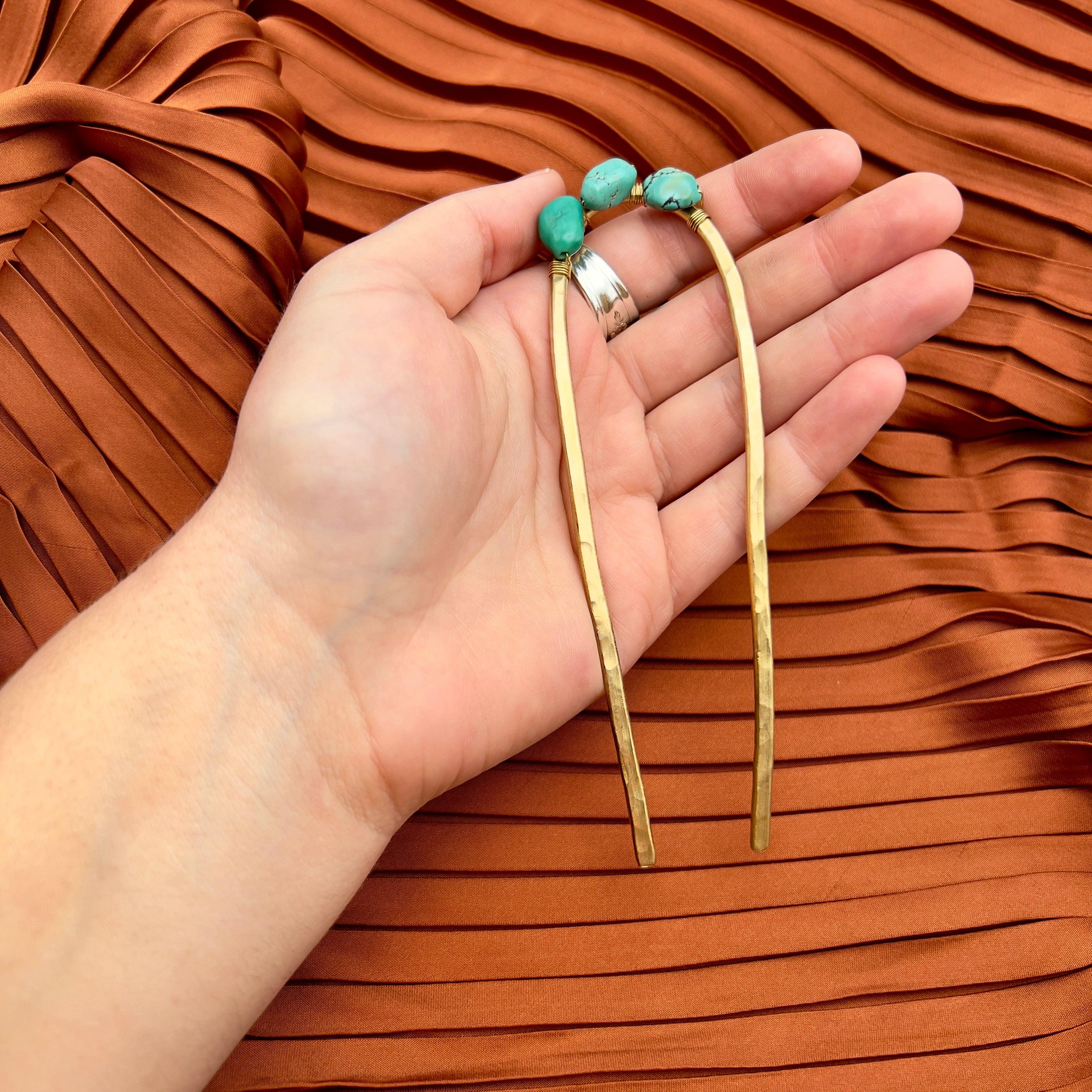 A hand holds the Fluff Hardware Turquoise Hair Fork, a gold-colored metal accessory topped with turquoise beads, set against a rust-colored pleated fabric background.