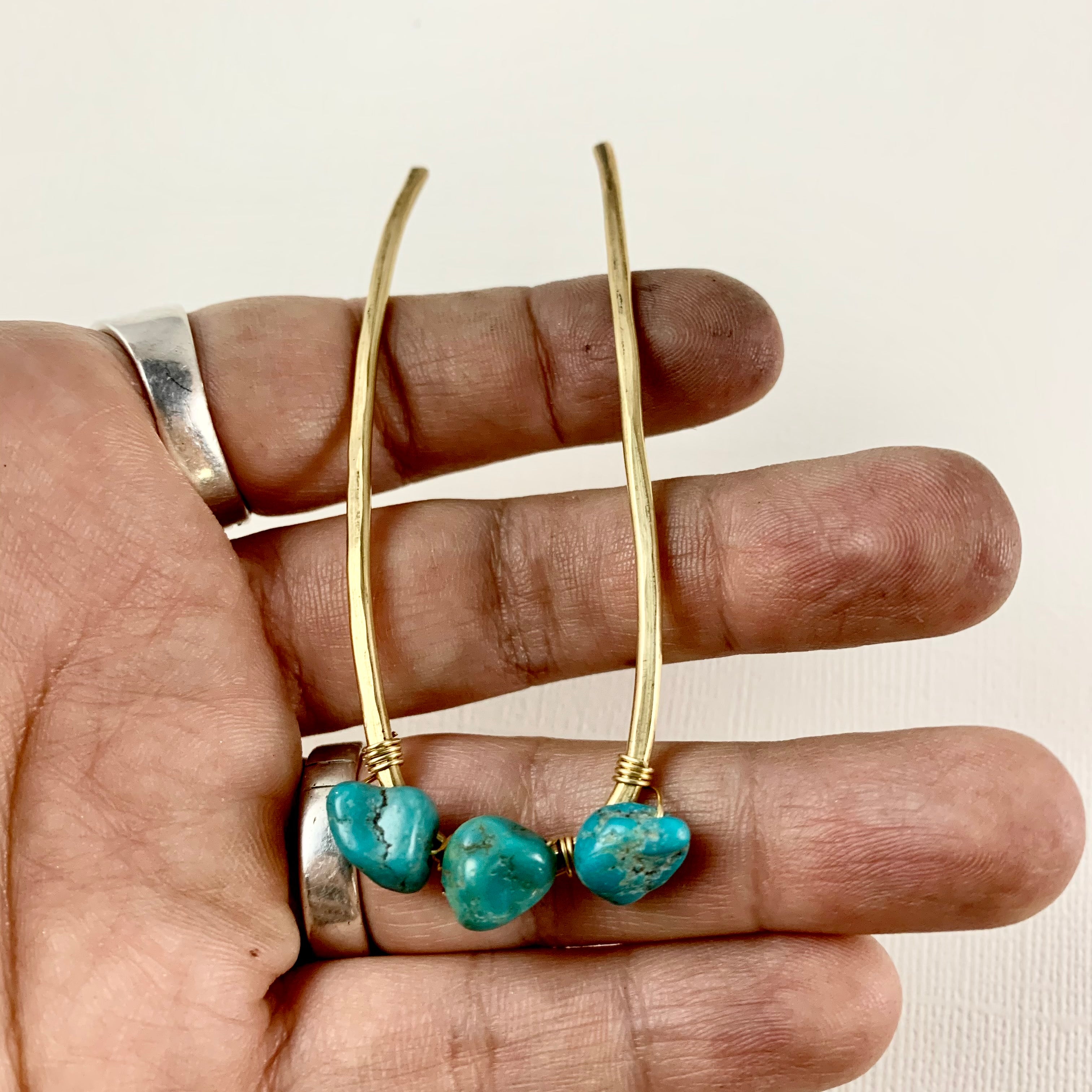 A hand holds two gold-colored Turquoise Hair Forks by Fluff Hardware, each with a small turquoise bead at the bottom. The person’s fingers, adorned with silver rings, are visible.