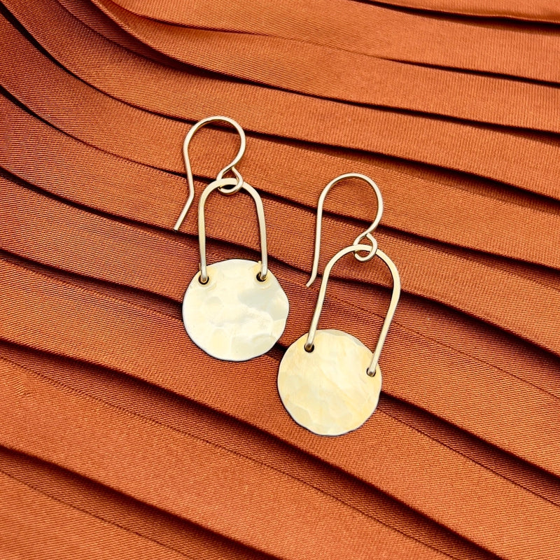 The Fallen Moon Earrings by Fluff Hardware are gold-toned, hammered metal dangles with circular discs, shown on pleated rust-orange fabric—ideal for those looking for stylish, hypoallergenic jewelry.