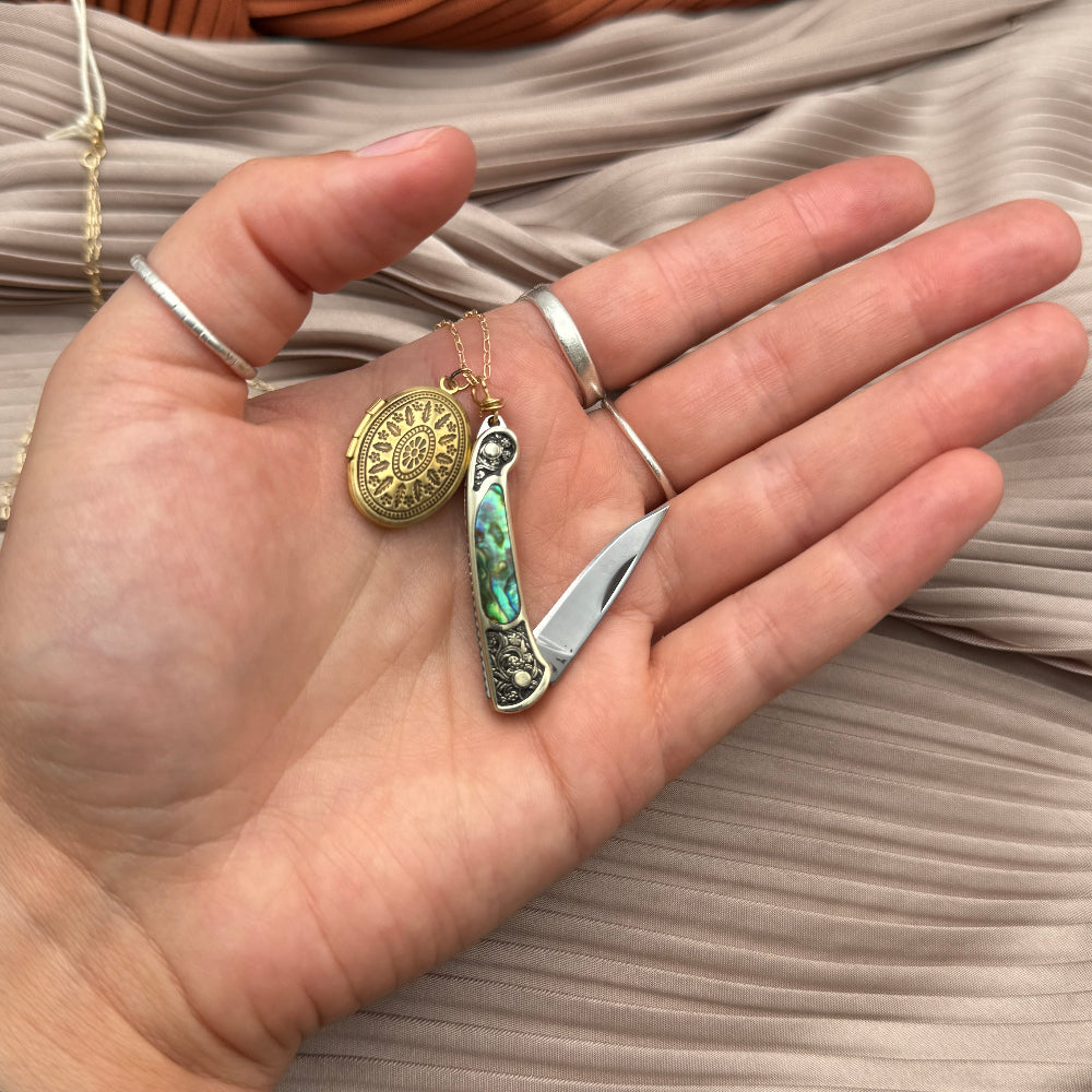 A hand displays the Fluff Hardware Keepsake Necklace—a gold oval locket with a sun design—next to a small pocket knife with abalone inlay, both on pleated taupe fabric.