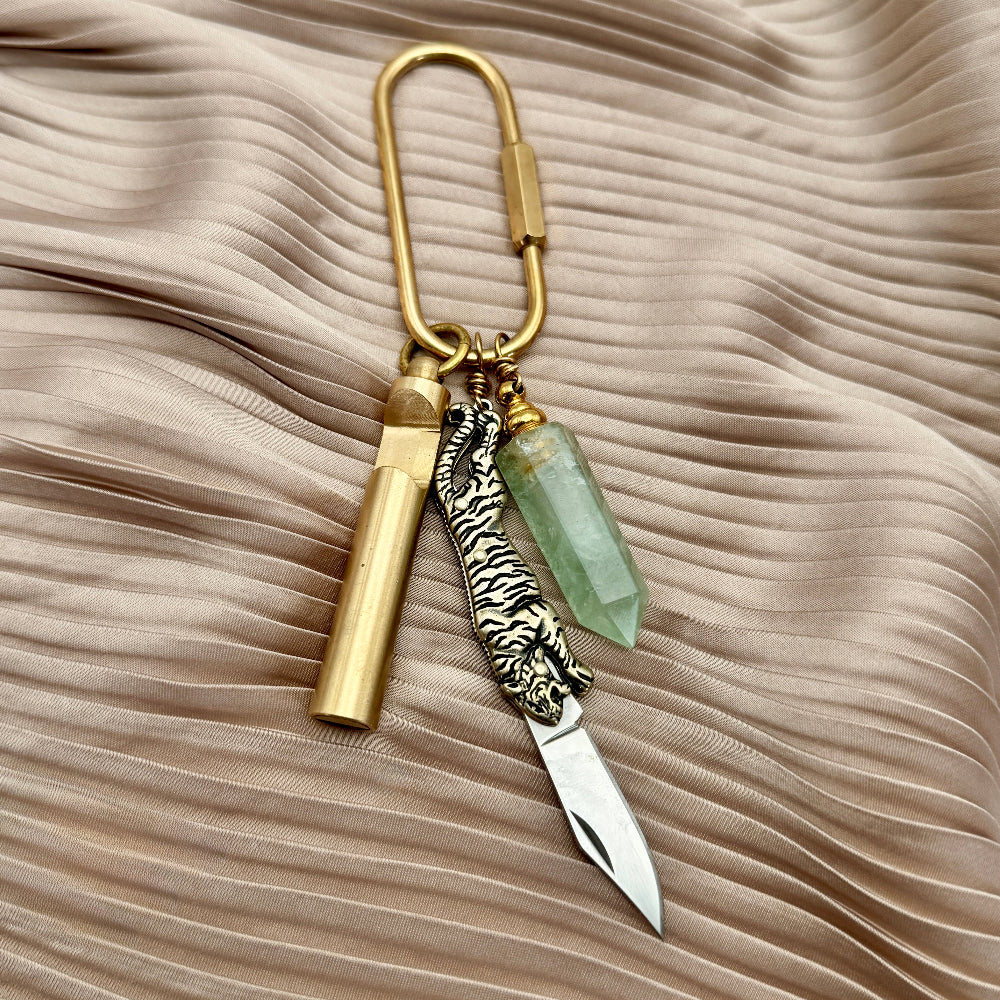 Keychain with gold cylindrical charm, silver tiger head charm, and green crystal on a beige fabric background