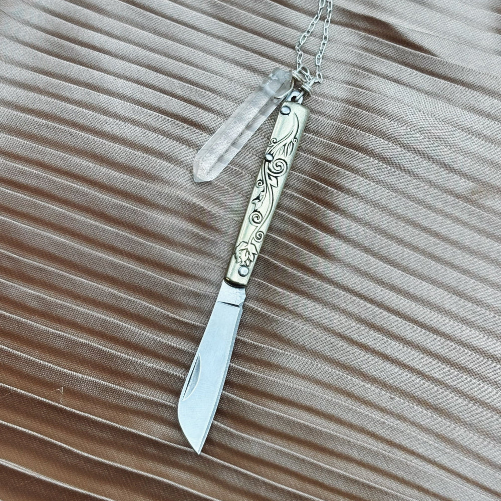 The Amelia Necklace by Fluff Hardware features a silver folding knife with an engraved handle and a quartz crystal pendant on a chain, shown against a textured beige surface.