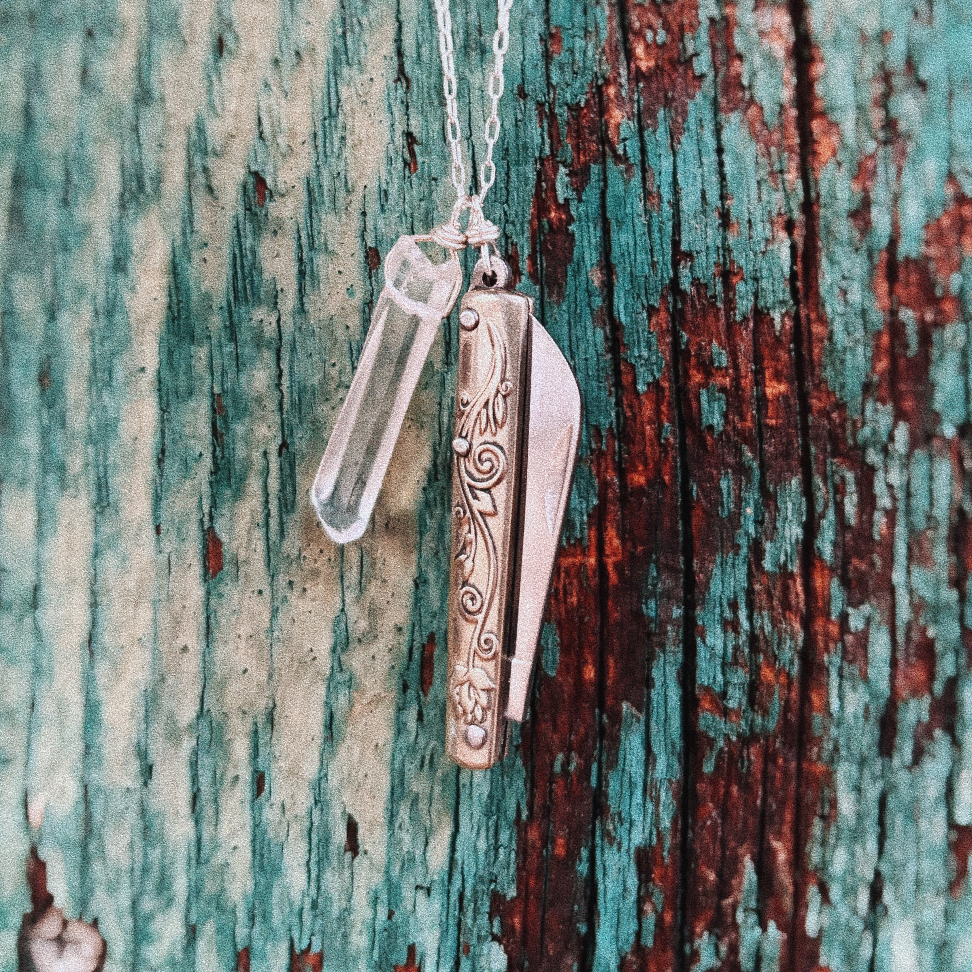 The Amelia Necklace by Fluff Hardware features a decorative pocketknife charm and a quartz crystal vial pendant, displayed on a weathered turquoise and brown wood background—ideal for fans of functional jewelry.