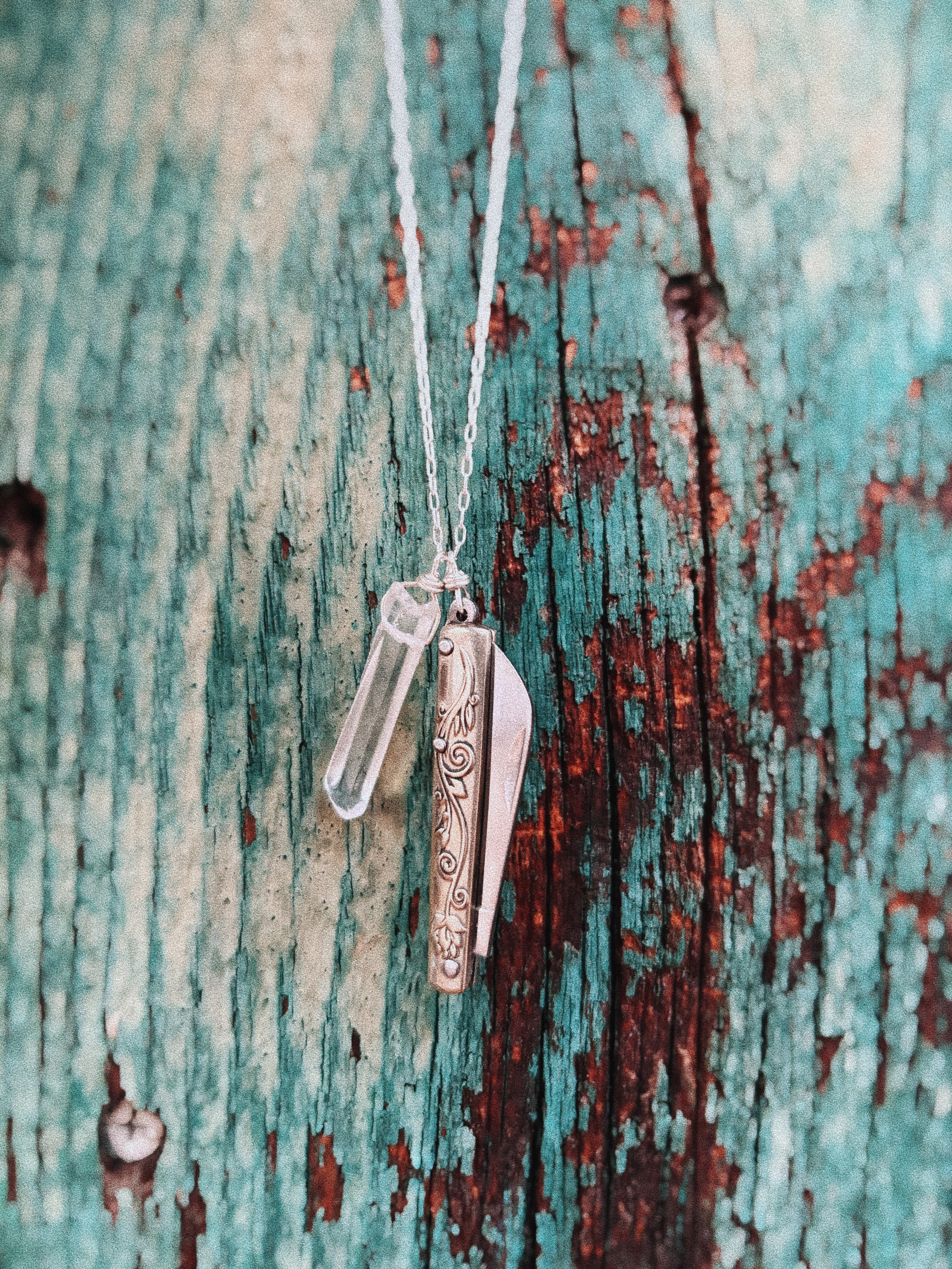 The Amelia Necklace by Fluff Hardware features a decorative pocketknife charm and a quartz crystal vial pendant, displayed on a weathered turquoise and brown wood background—ideal for fans of functional jewelry.