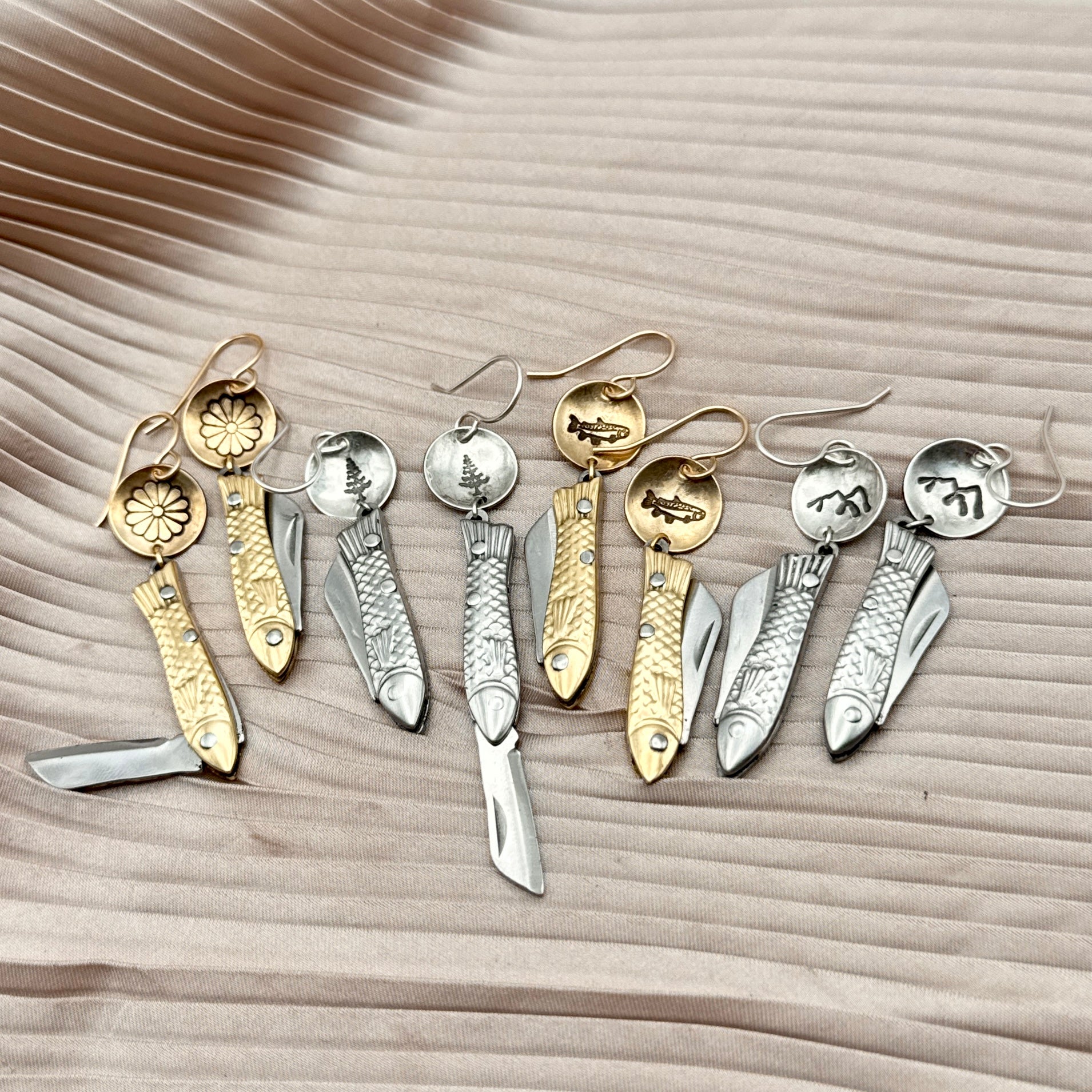 Four pairs of Backcountry Feeesh Earrings by Fluff Hardware in gold and sterling silver tones, each featuring a unique fish-shaped mini pocket knife pendant,  displayed on pleated beige fabric, showcasing flower, tree, fish, and mountain stamp