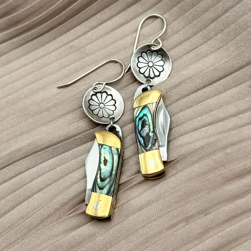 Fluff Hardwares Backcountry Earrings (Abalone) feature round silver discs stamped with flower and an abalone inlay pocket knife pendant earrings with gold accents, blending artistry and function atop beige pleated fabric.