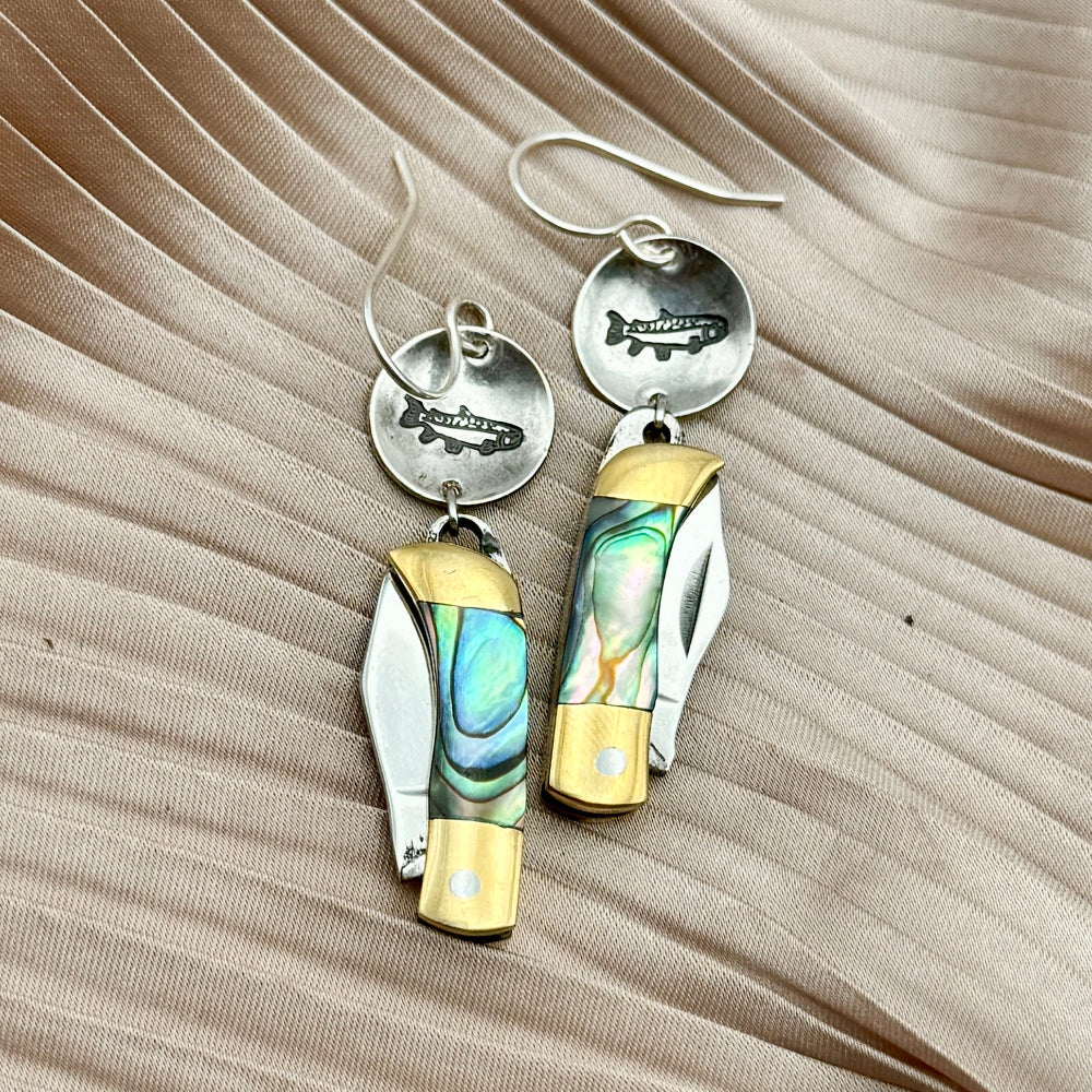 Fluff Hardwares Backcountry Earrings (Abalone) feature round silver discs stamped with fish and an abalone inlay pocket knife pendant earrings with gold accents, blending artistry and function atop beige pleated fabric.