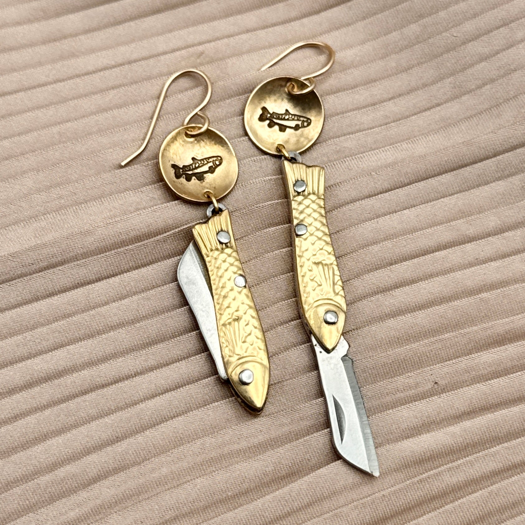 The Fluff Hardware Backcountry Feeesh Earrings feature detailed sterling silver fish designs with a fish stamped round metal piece, shown on beige textured fabric—an elegant blend of artistry and functionality.