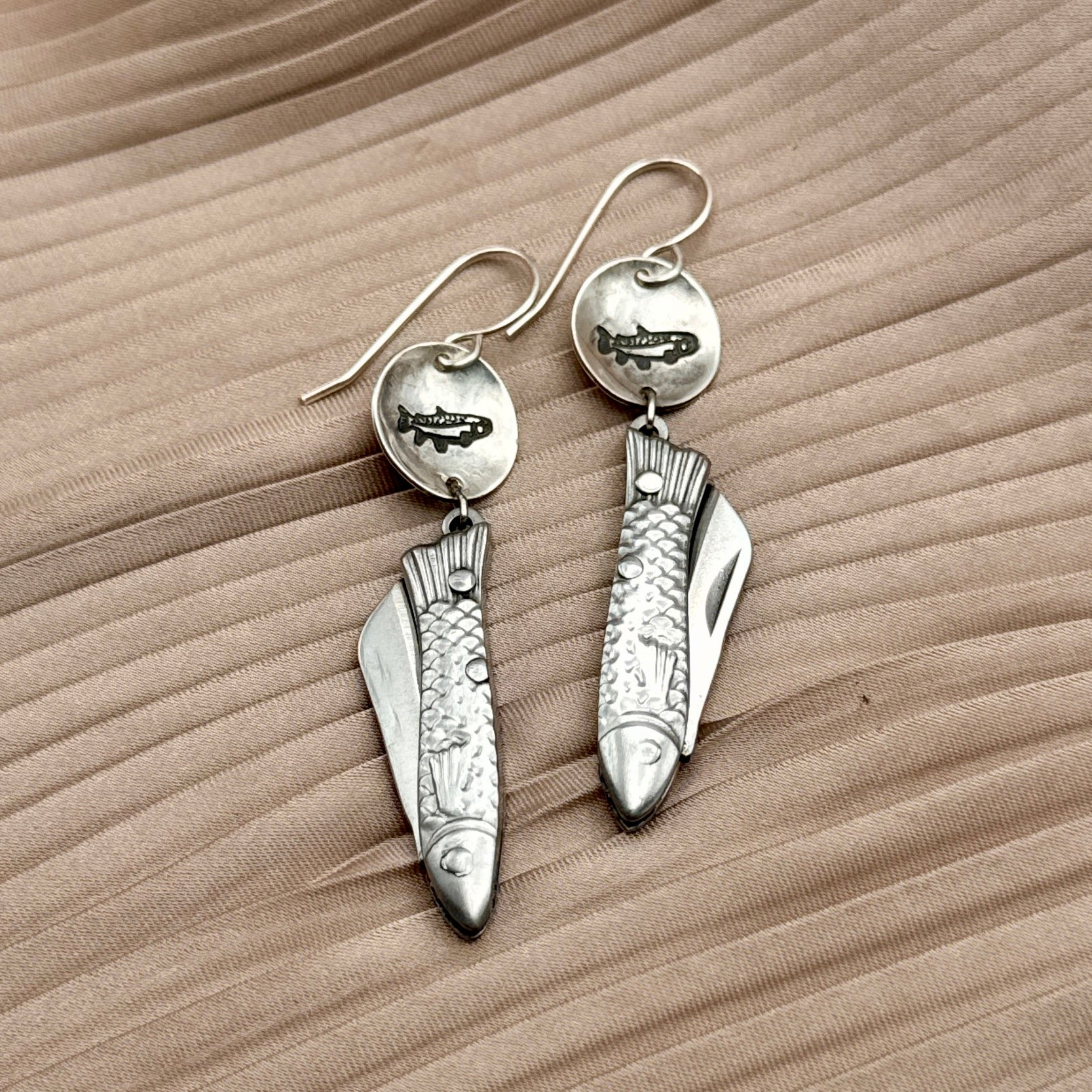 The Fluff Hardware Backcountry Feeesh Earrings feature detailed sterling silver fish designs with a fish stamped round metal piece, shown on beige textured fabric—an elegant blend of artistry and functionality.