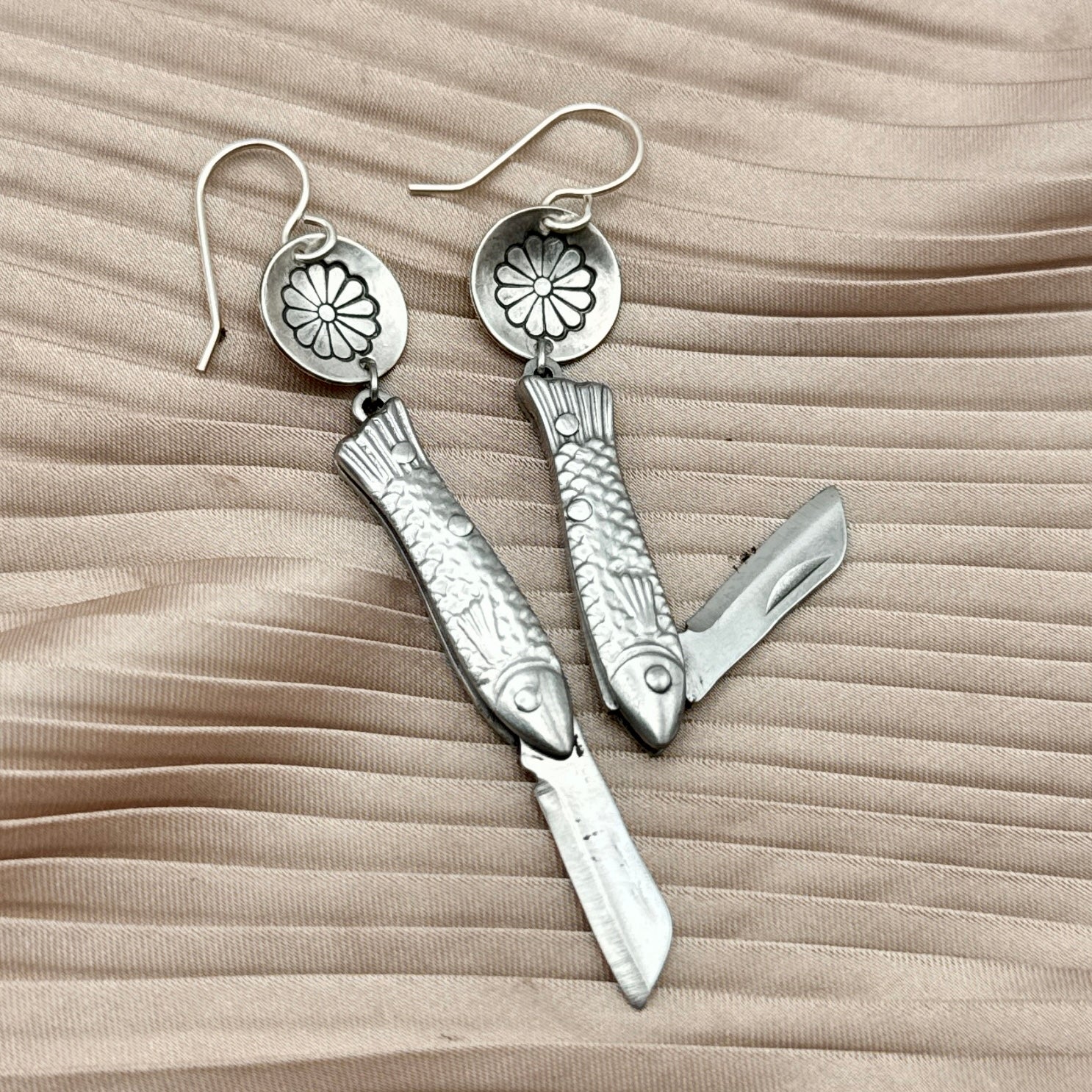 The Backcountry Feeesh Earrings by Fluff Hardware are silver fish-shaped pocket knife earrings with engraved scales; tiny knife blades from charm. These functional hook-wire earrings are shown against pleated beige fabric.