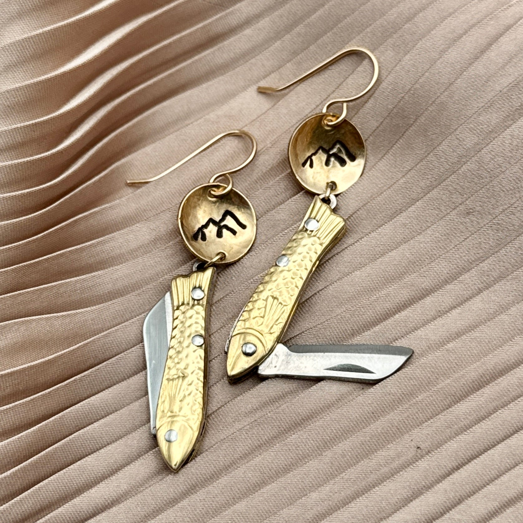 The Backcountry Feeesh Earrings by Fluff Hardware feature gold fish-shaped pendants with circular, engraved mountain tops, blending artistry and everyday elegance. Displayed on textured beige fabric.