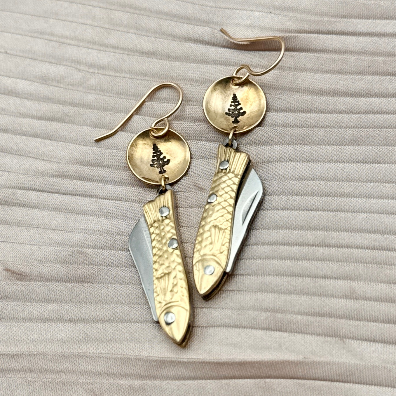 The Backcountry Feeesh Earrings by Fluff Hardware are silver or gold earrings with miniature fish pocketknife charms, each hanging from a round gold disc engraved with a pine tree, displayed on textured beige fabric.