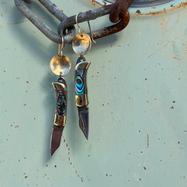 A pair of earrings featuring an abalone shell inlay on a small metallic utility tool pocketknife, with one earring in part open position to display the blade, hanging from a silver ear wire.