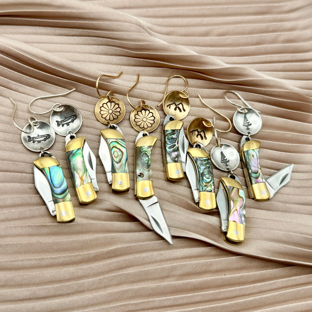 Fluff Hardware’s Backcountry Earrings (Abalone) feature miniature folding knives with abalone inlay and gold accents, paired with stamped round metal charms, beautifully displayed on beige pleated fabric.