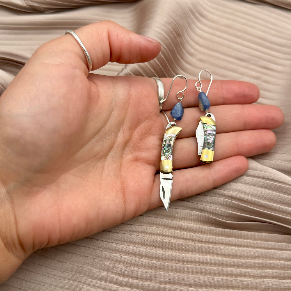 Hand holding two abalone mini pocket knife earrings with a kyanite bead hanging above on a neutral background