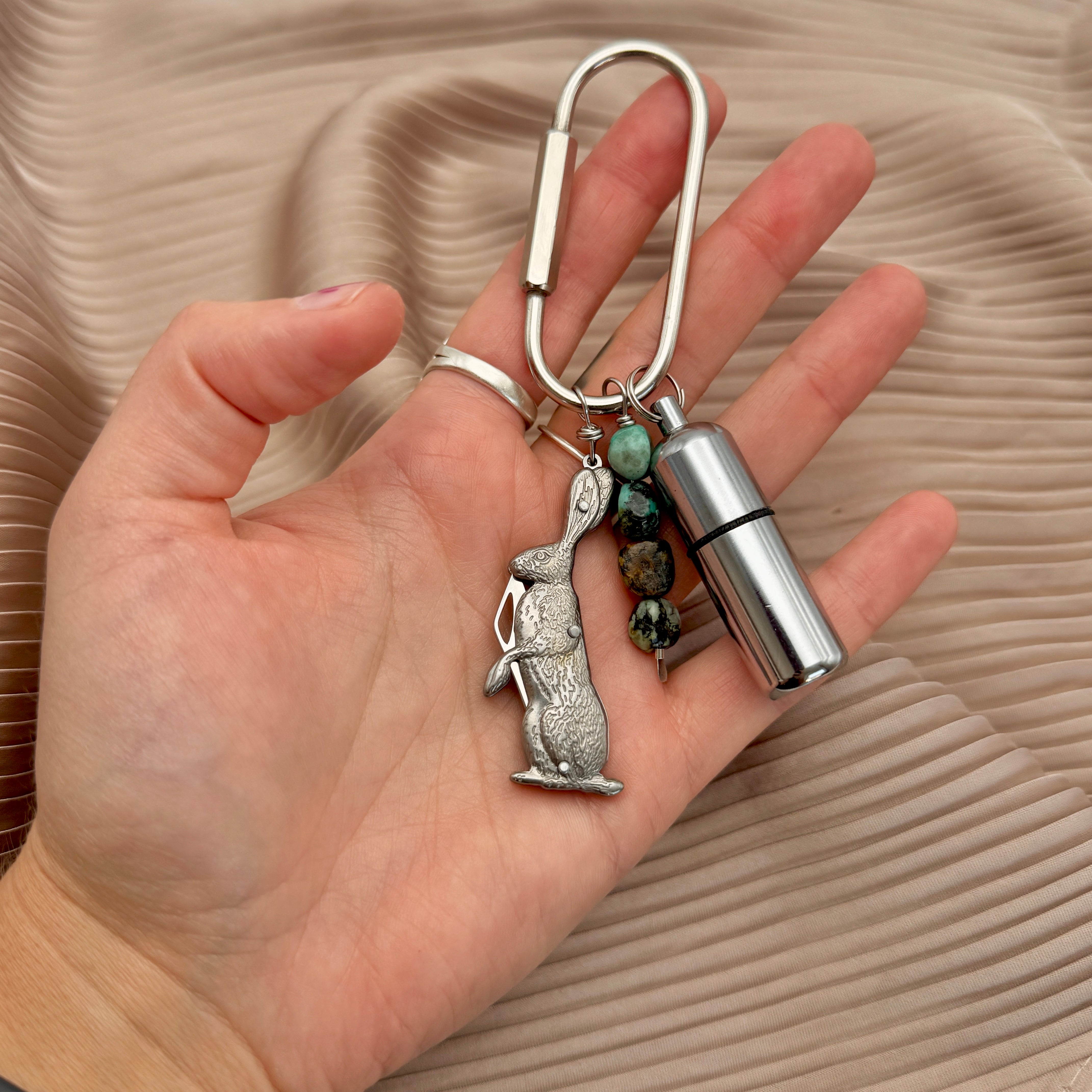 A hand holding a silver carabiner keychain with a rabbit charm, turquoise beads, and a silver cylindrical lighter, set against a pleated beige fabric background.