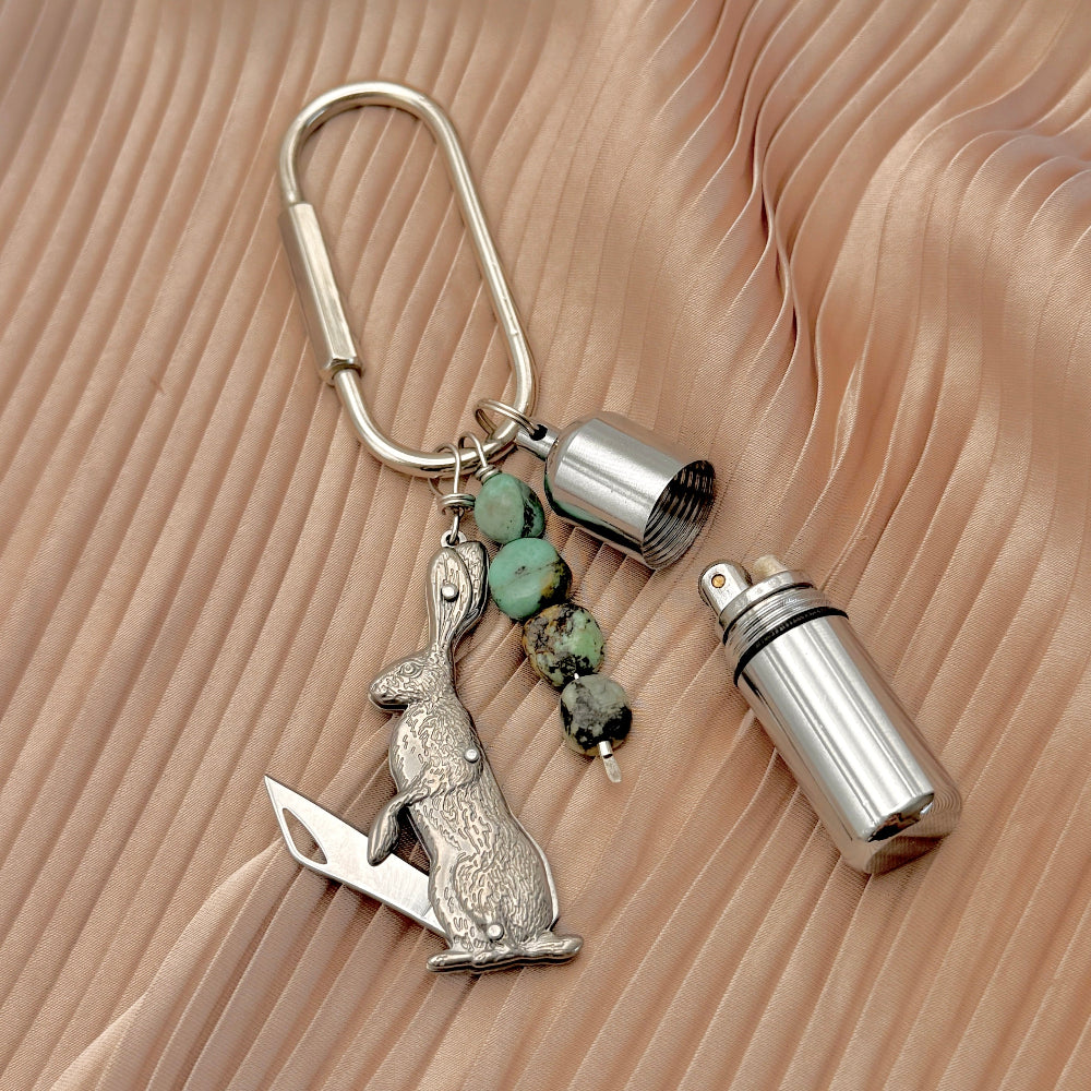 A silver metal keychain featuring a rabbit-shaped tool, a small cylindrical metal lighter, and four turquoise beads, displayed on a beige pleated fabric background.