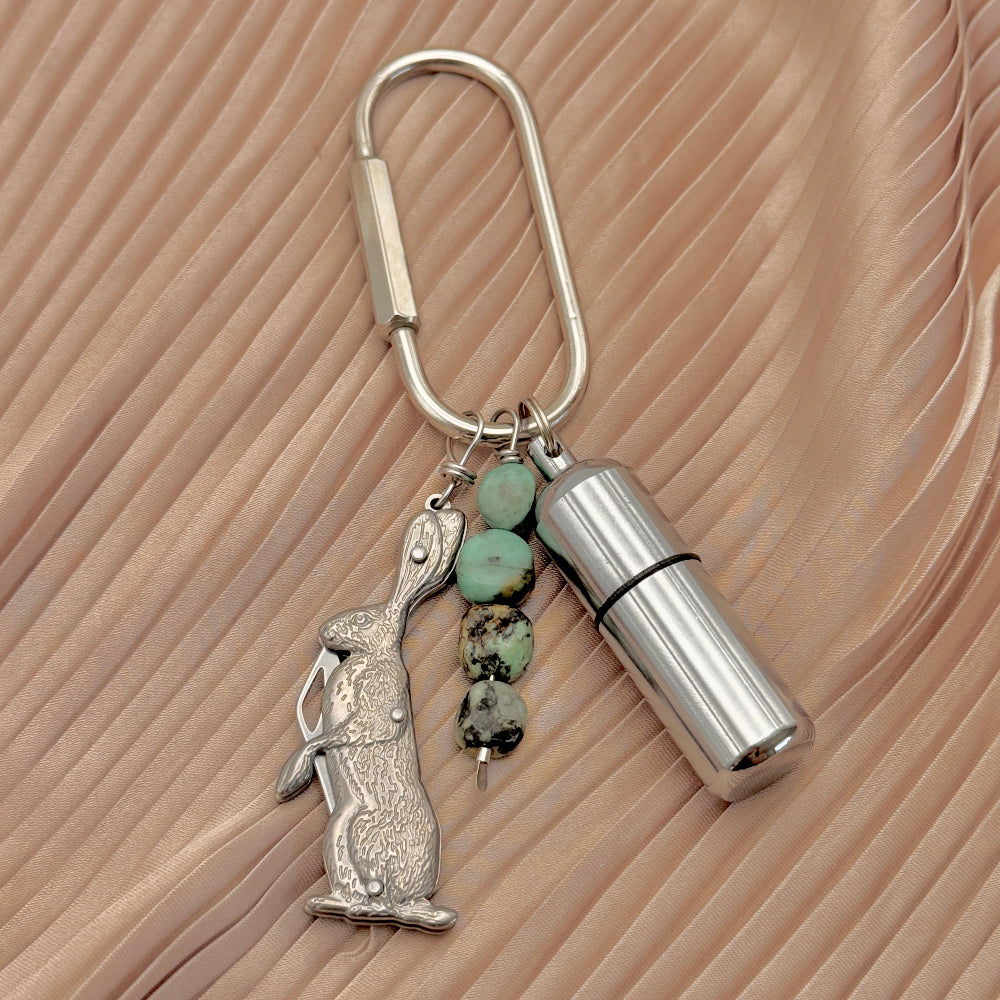 A silver carabiner keychain featuring a rabbit charm, a cylindrical metal lighter, and four turquoise beads, displayed on a textured beige background.