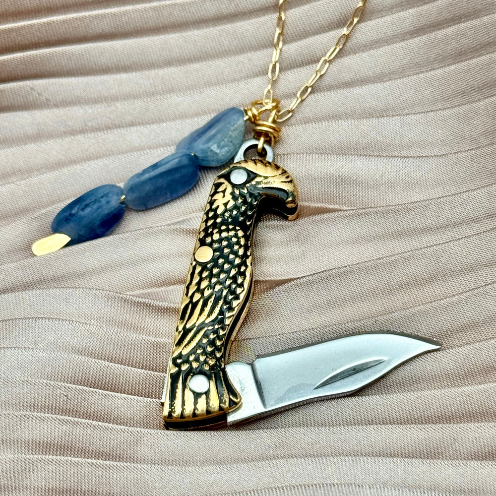 Necklace with an eagle-shaped pocket knife pendant with the knife open 
and blue kyanite beads on a beige fabric background