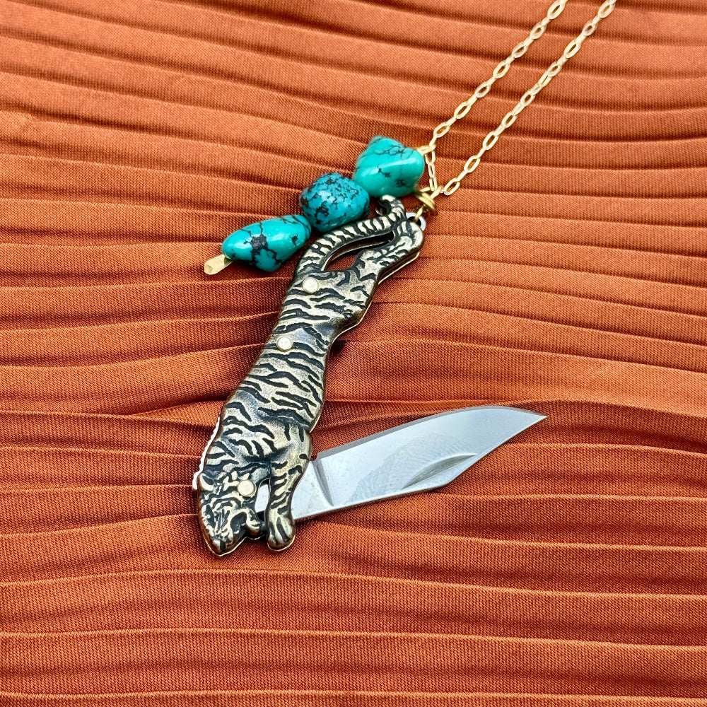 Tiger-shaped pocket knife with turquoise beads on a gold chain against an orange fabric background