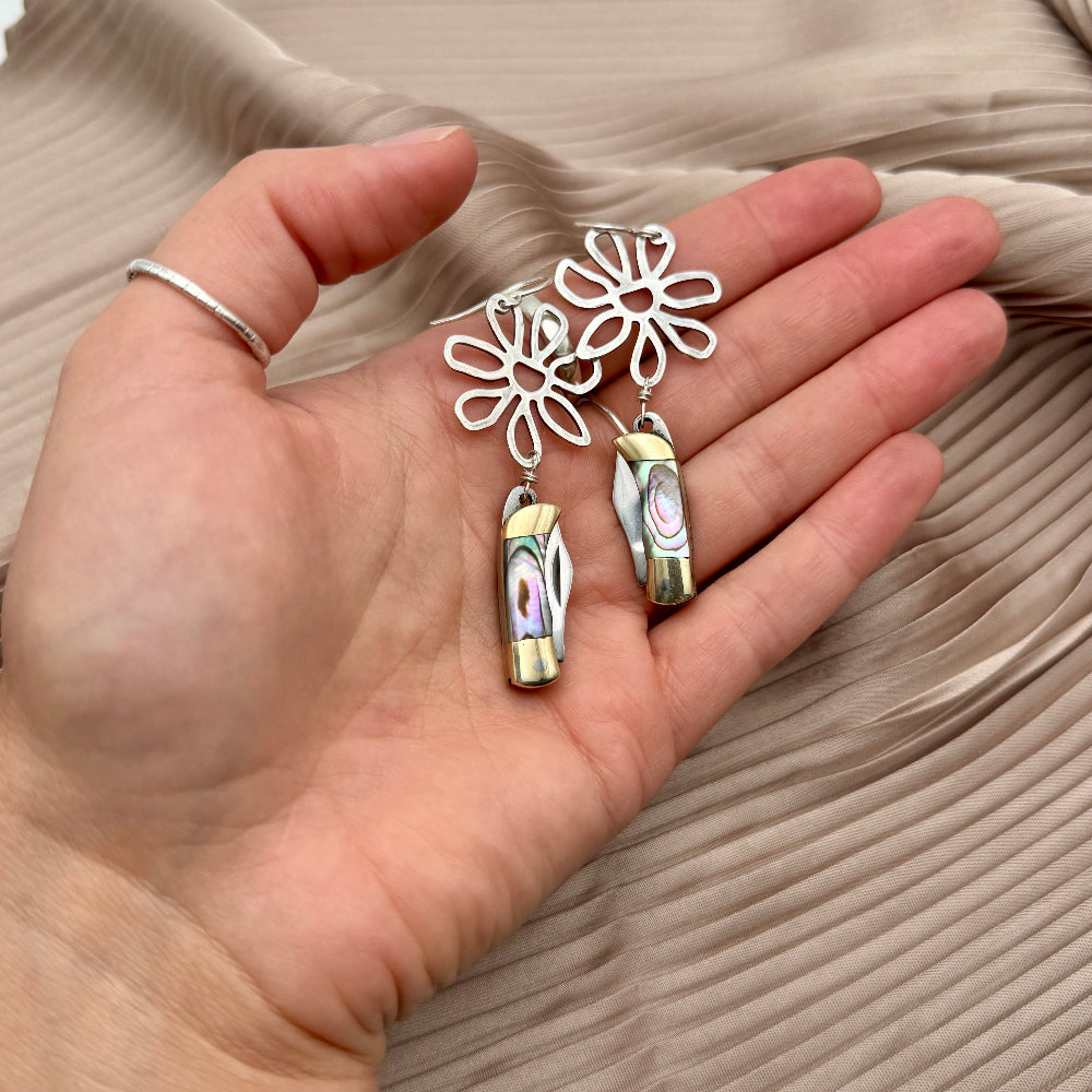 Flower pendant with miniature pocketknives hanging off of silver ear wire held in a hand over a drape