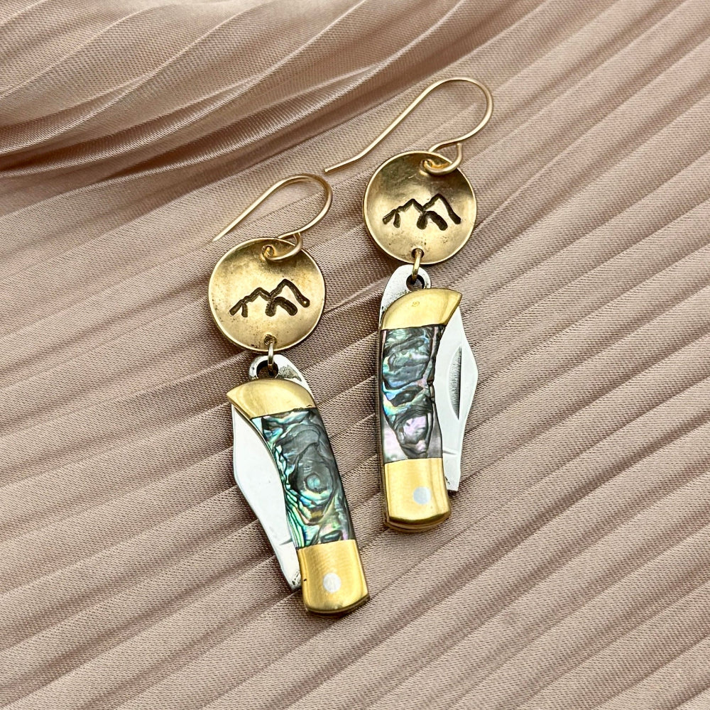 Fluff Hardwares Backcountry Earrings (Abalone) feature round silver discs stamped with mountain and an abalone inlay pocket knife pendant earrings with gold accents, blending artistry and function atop beige pleated fabric.